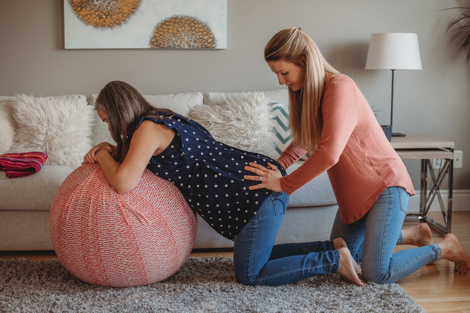 Pregnant woman leaning on pink exercise ball while supportive companion massages her back during labor at home.
