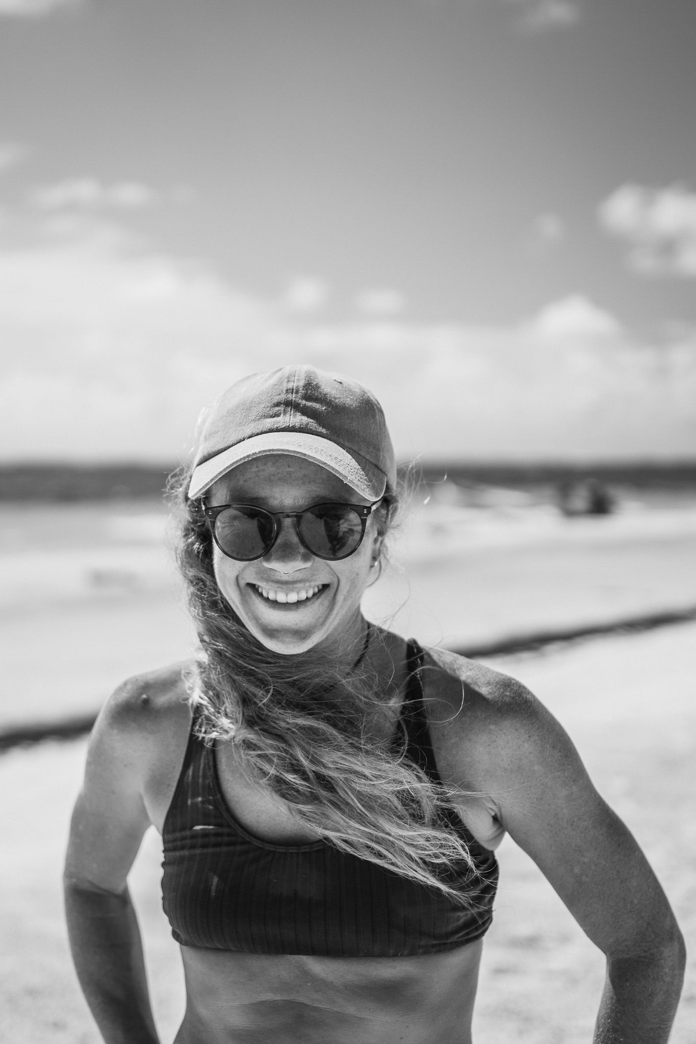 Black and white photo of a smiling woman in sunglasses and cap on a windy beach wearing a sports bra.