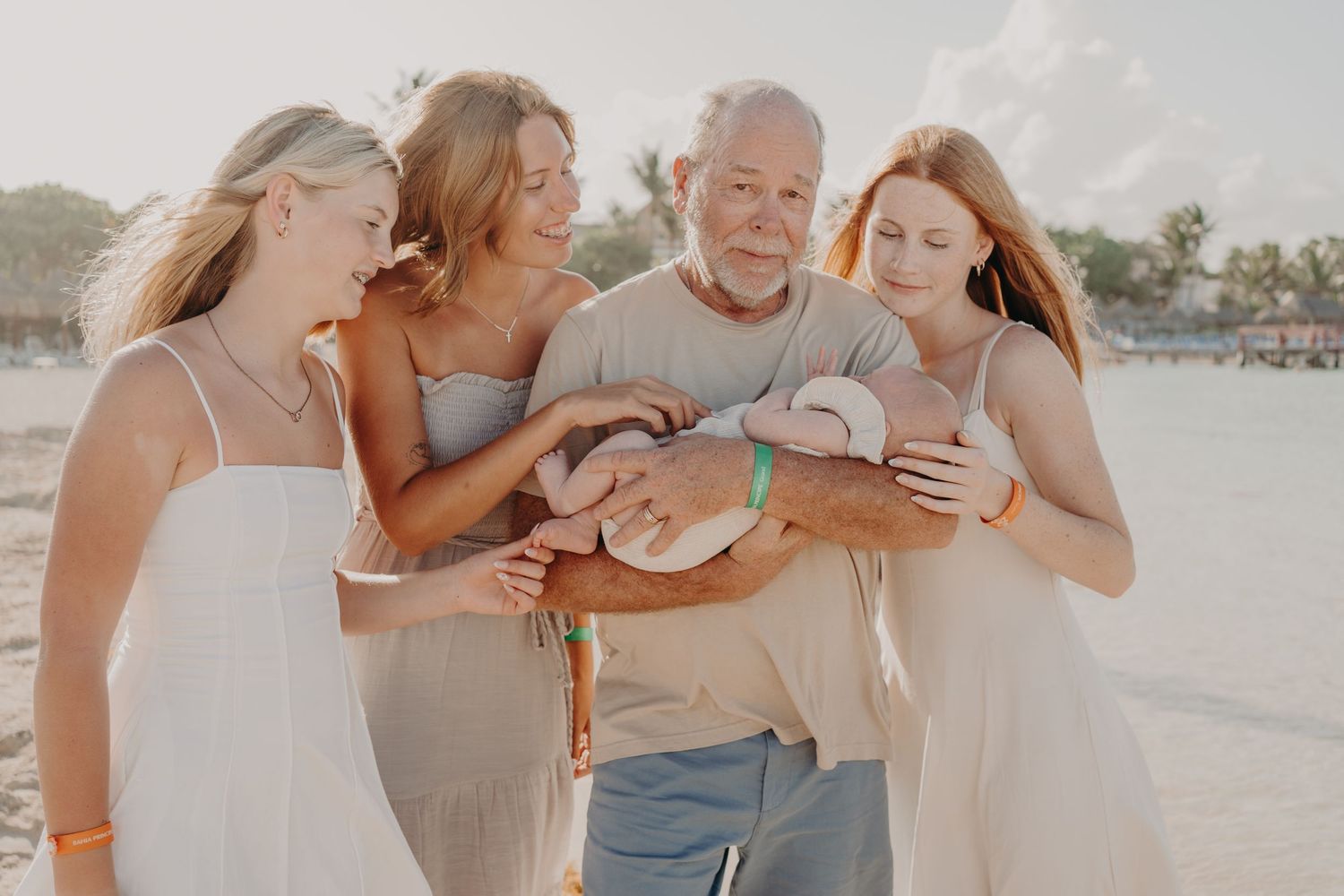Family gathered on beach admiring newborn baby, smiling adults in light summer attire with ocean backdrop.