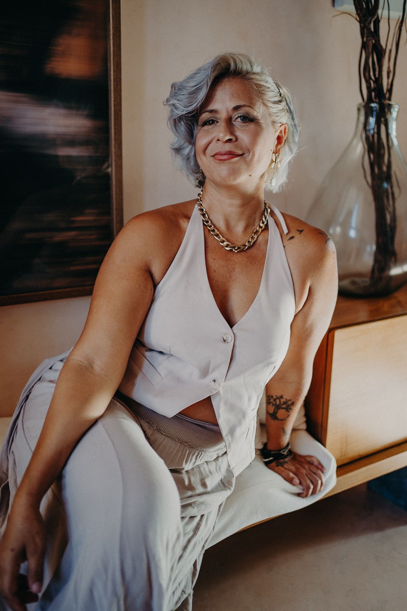 Woman with silver hair sitting confidently in white halter outfit with gold chain necklace and tattoos.