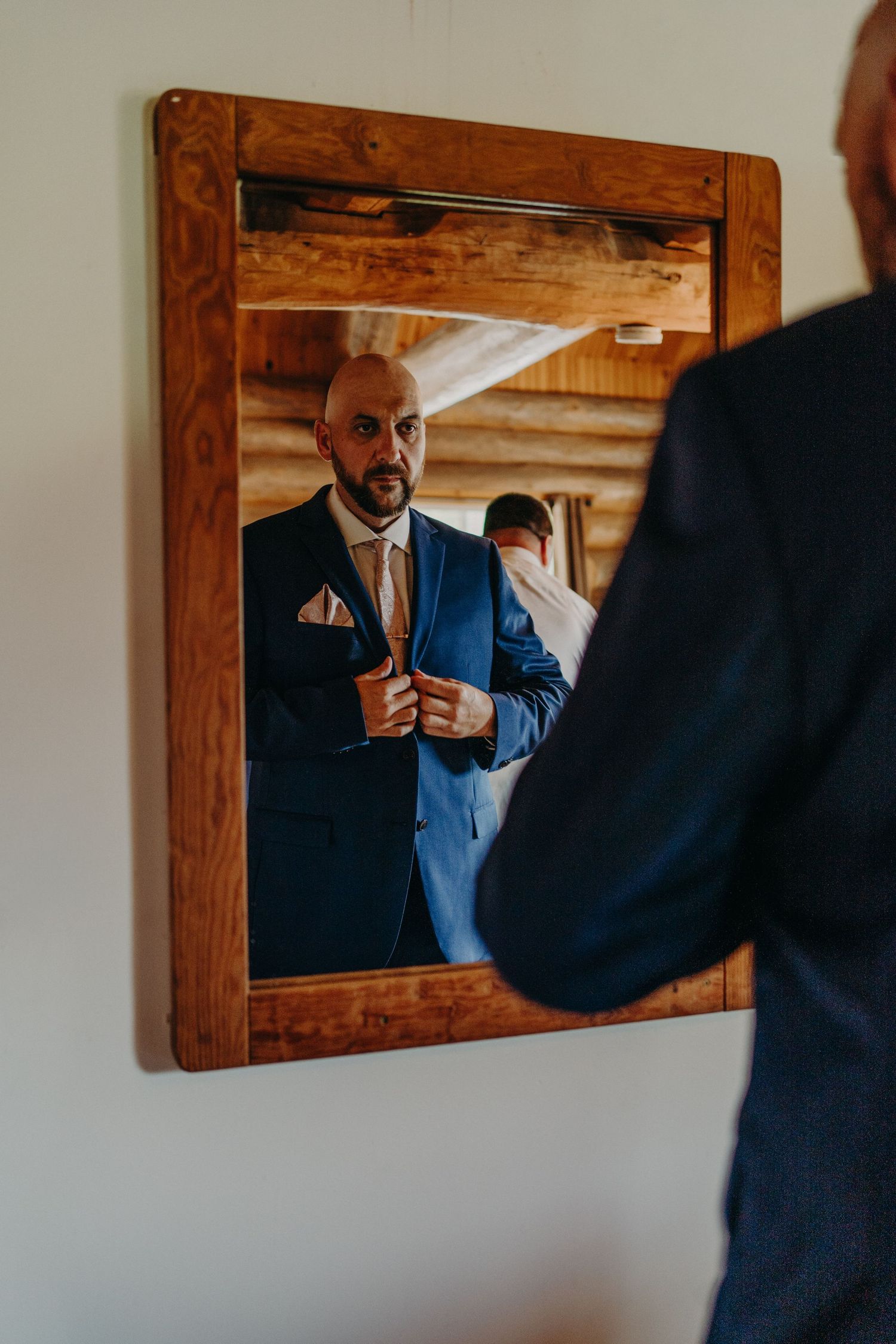 Groom in navy blue suit adjusting pocket square while viewing reflection in rustic wooden mirror.