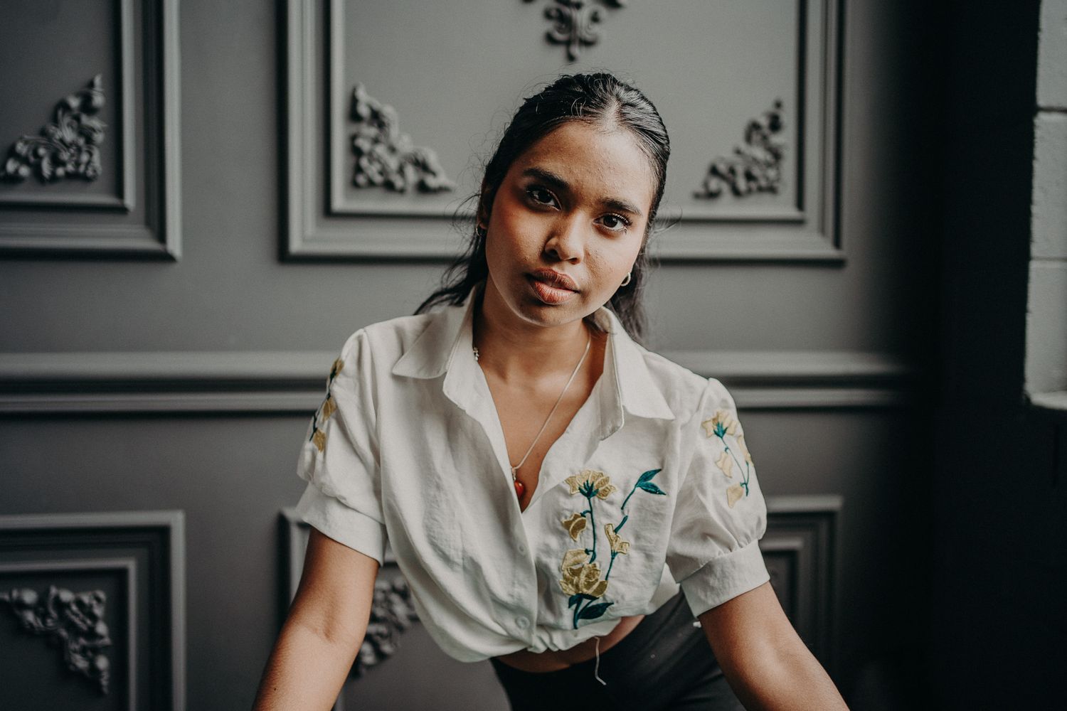 Young woman in embroidered white shirt poses thoughtfully in elegant gray interior setting.