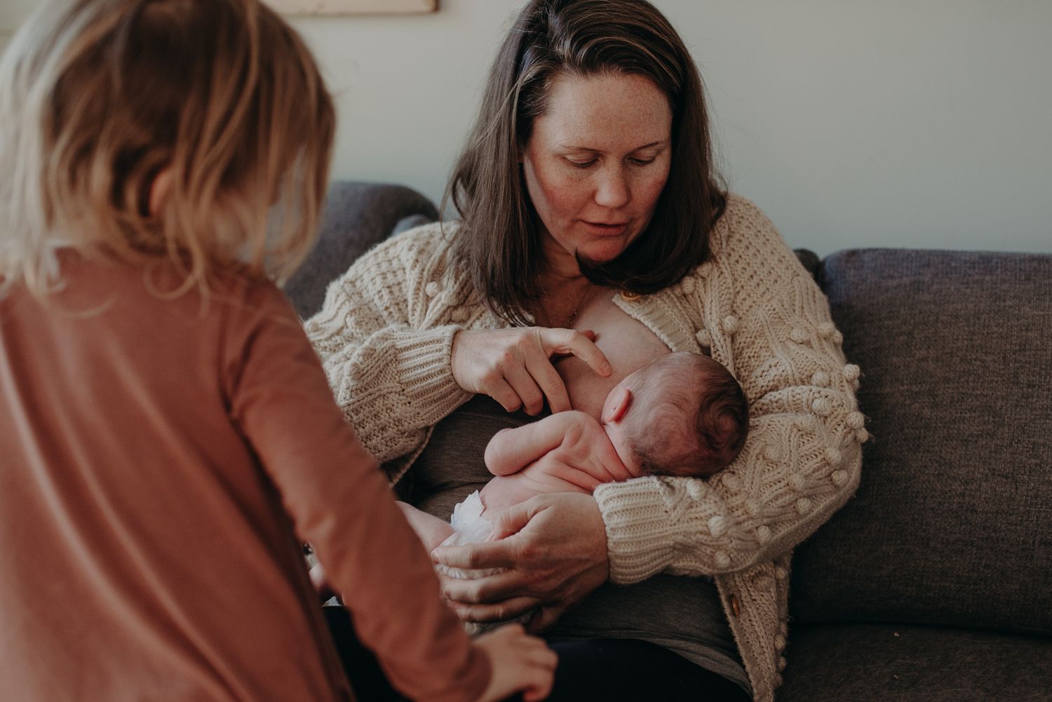 A mother sits on a couch breastfeeding her newborn baby while an older child stands nearby watching.