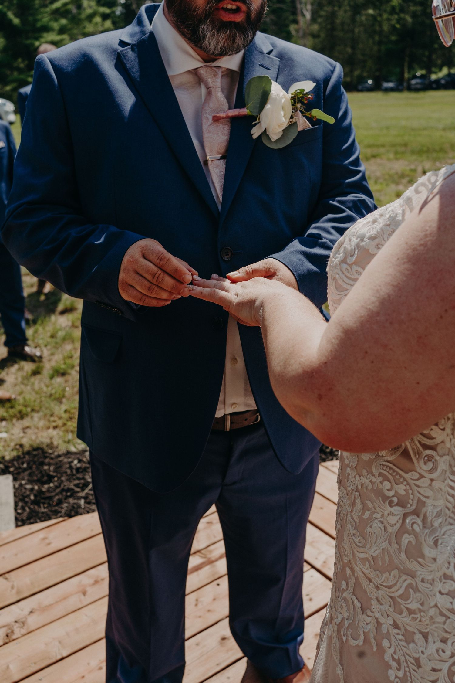 Groom in navy suit placing wedding ring on bride's finger during outdoor ceremony on wood deck.