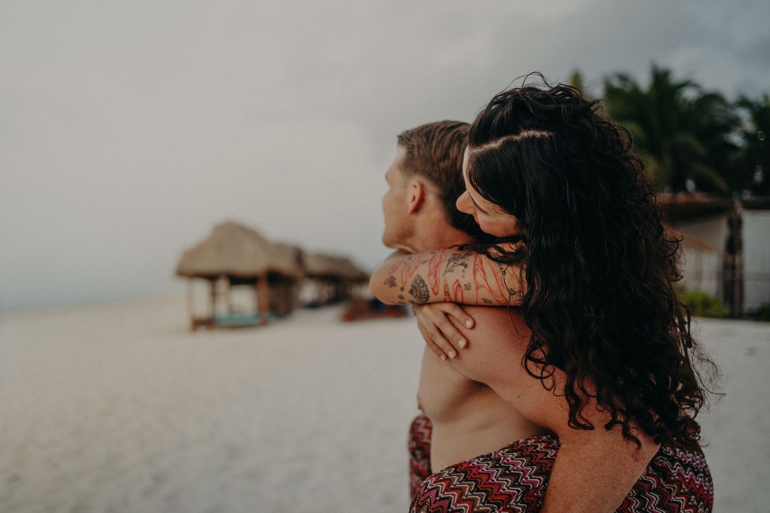 Couple embracing on a quiet tropical beach at dusk, with a woman's arms wrapped around a man and thatched palapa huts and palm trees behind.