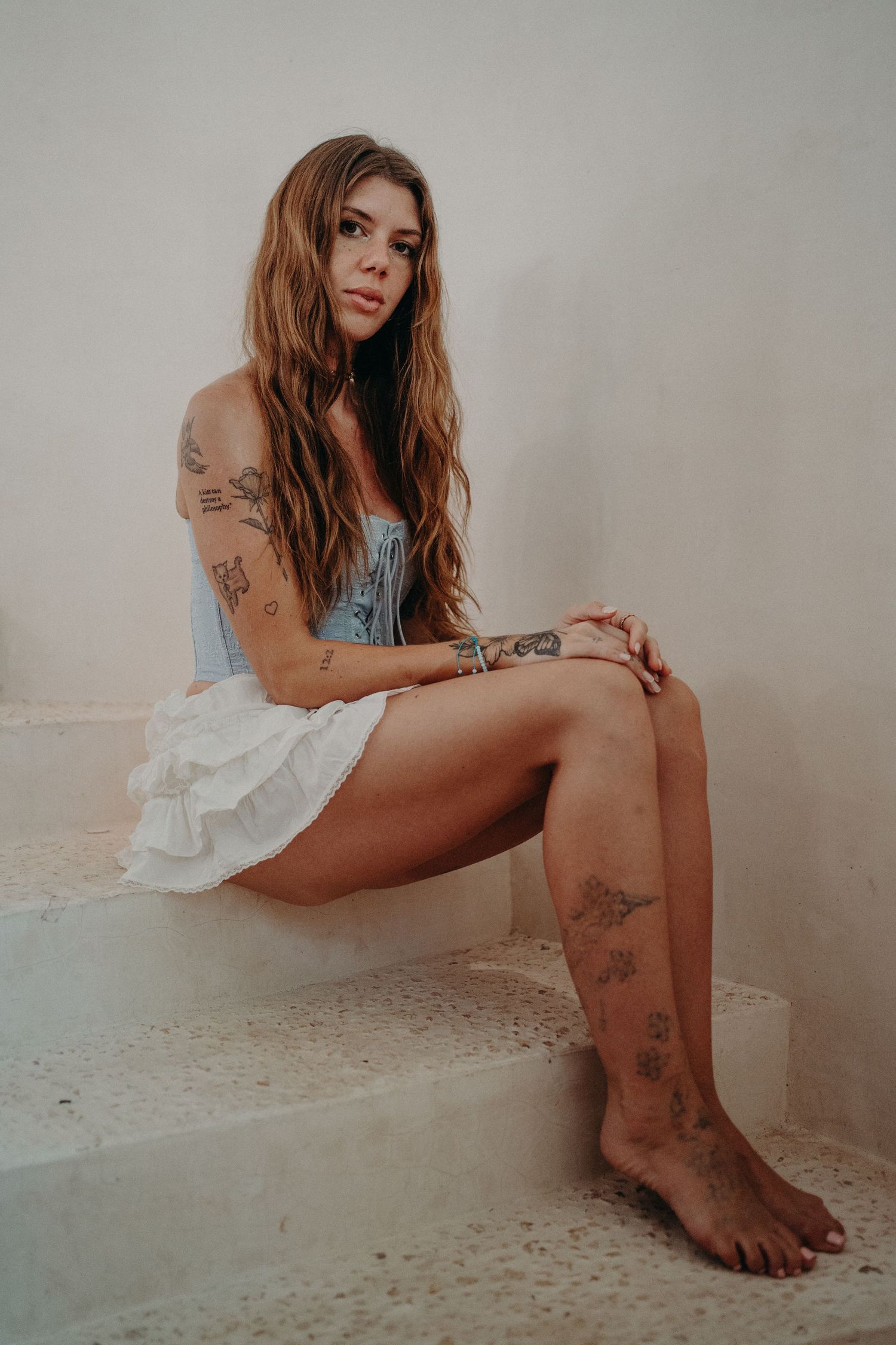Tattooed woman with long auburn hair in white mini dress sitting barefoot on white stone steps.