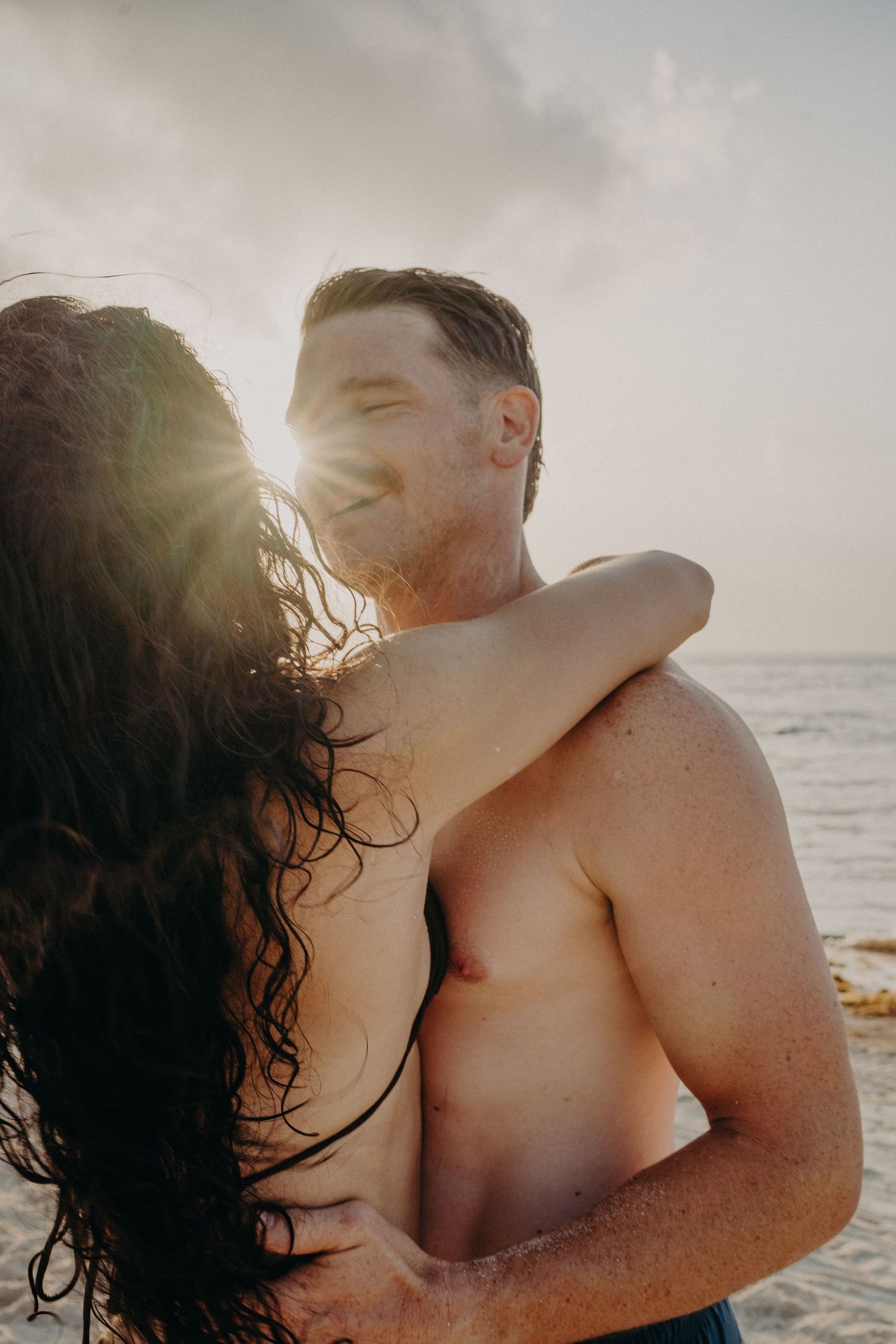 Couple embracing on a beach at sunset, shirtless man smiling with golden sunlight glowing behind them.