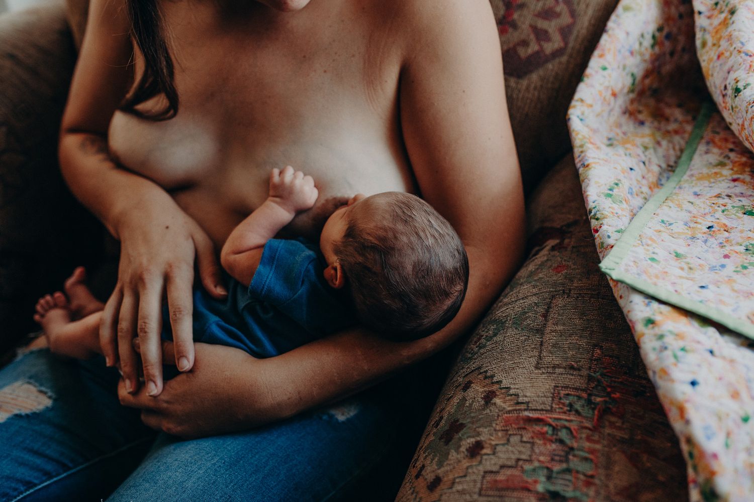 Mother breastfeeding newborn baby on a couch with floral cushions in a warm home setting.