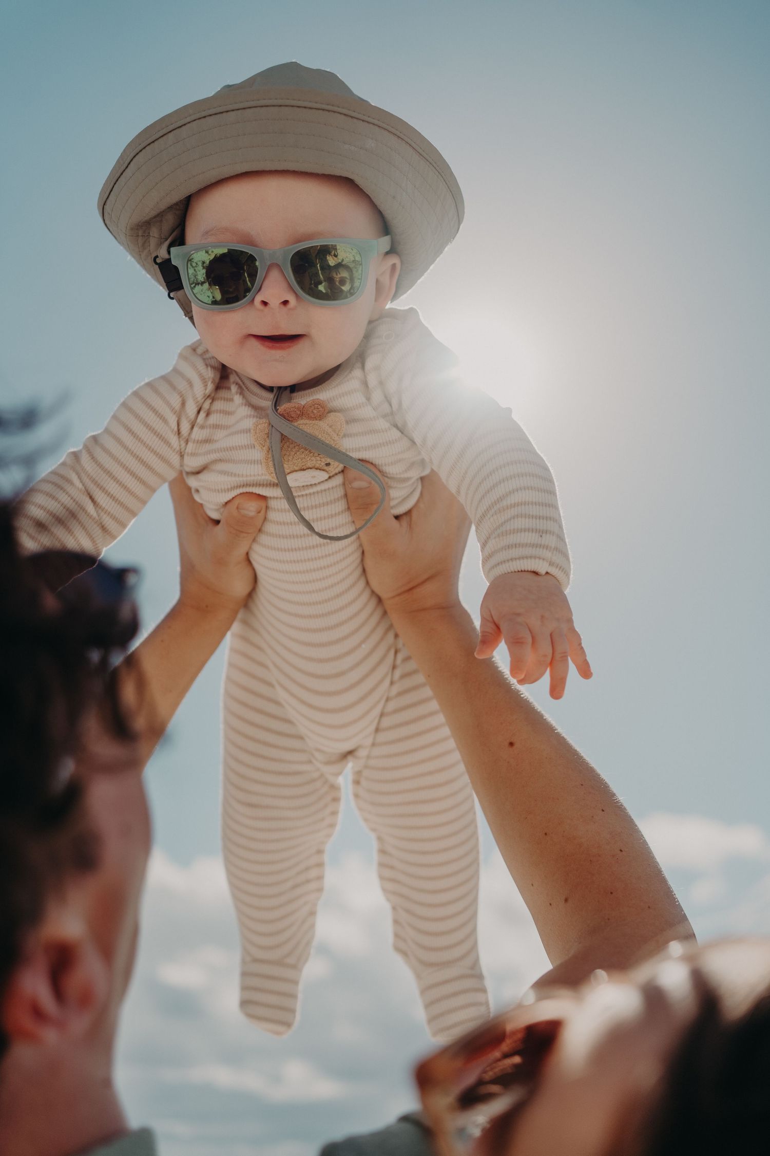 Adorable baby in sunglasses and hat held up by parents against bright sunny sky.