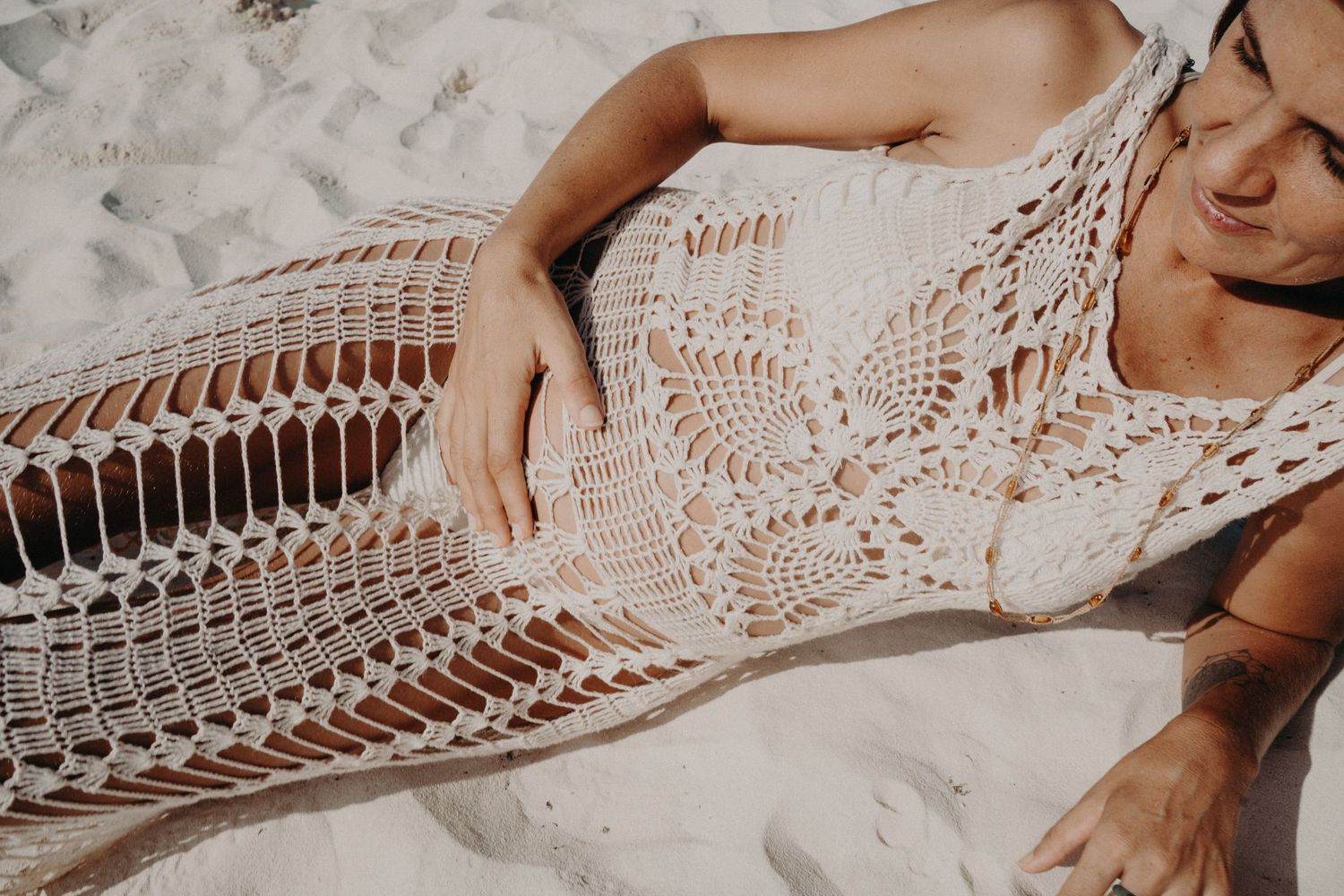 Woman reclining on white sand wearing a white crochet beach dress with intricate openwork lace details, perfect boho summer style.