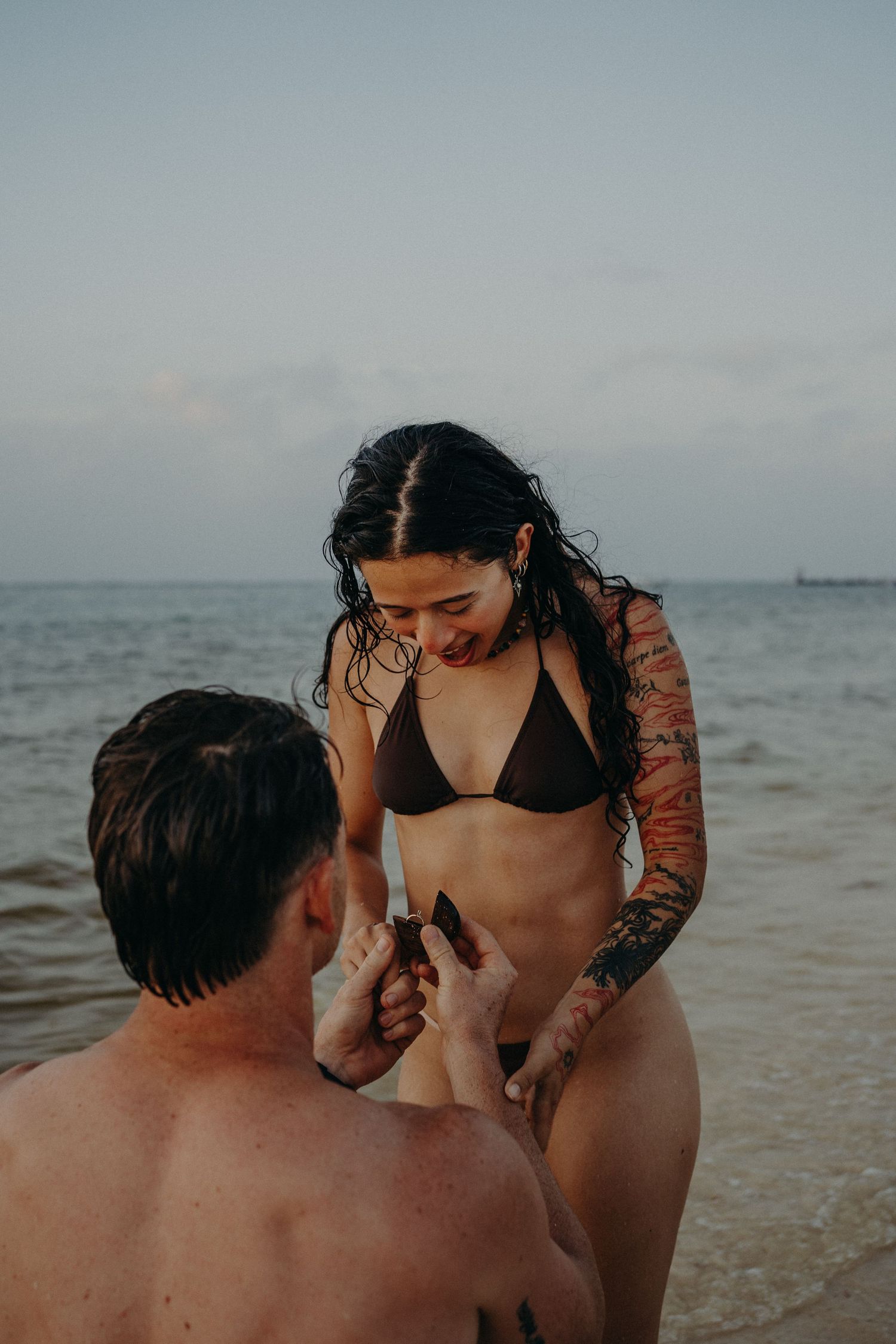 A man kneels in shallow ocean water holding a ring box as a smiling woman in a bikini looks down during a beach proposal.