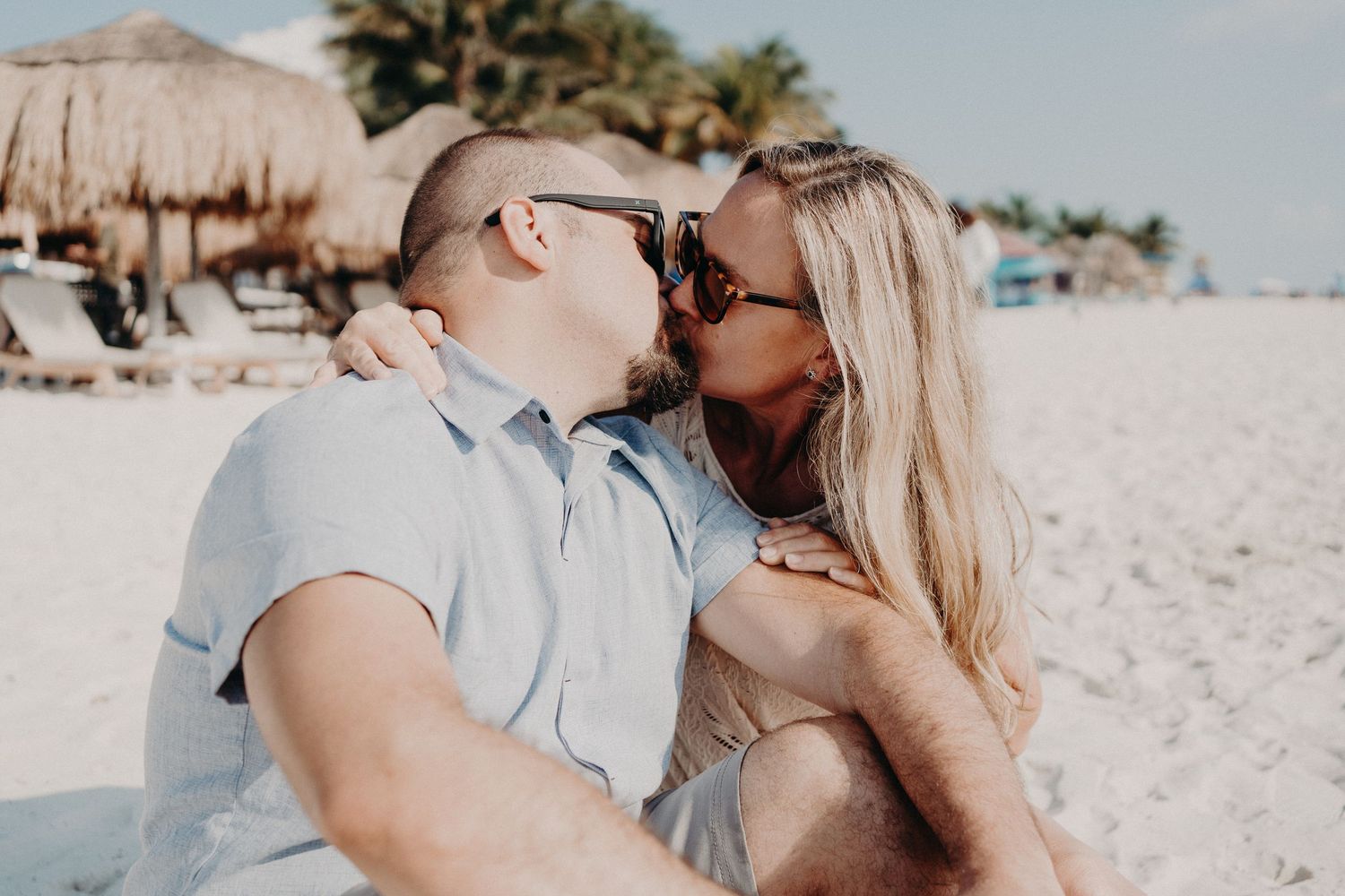 Couple kissing on a white sand beach with palm trees, both wearing sunglasses, romantic tropical vacation moment.
