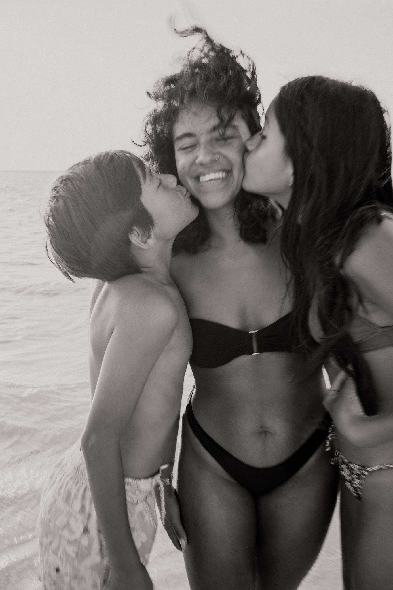 Three women in swimwear share joyful kisses on the cheek at the beach in a black and white photo.