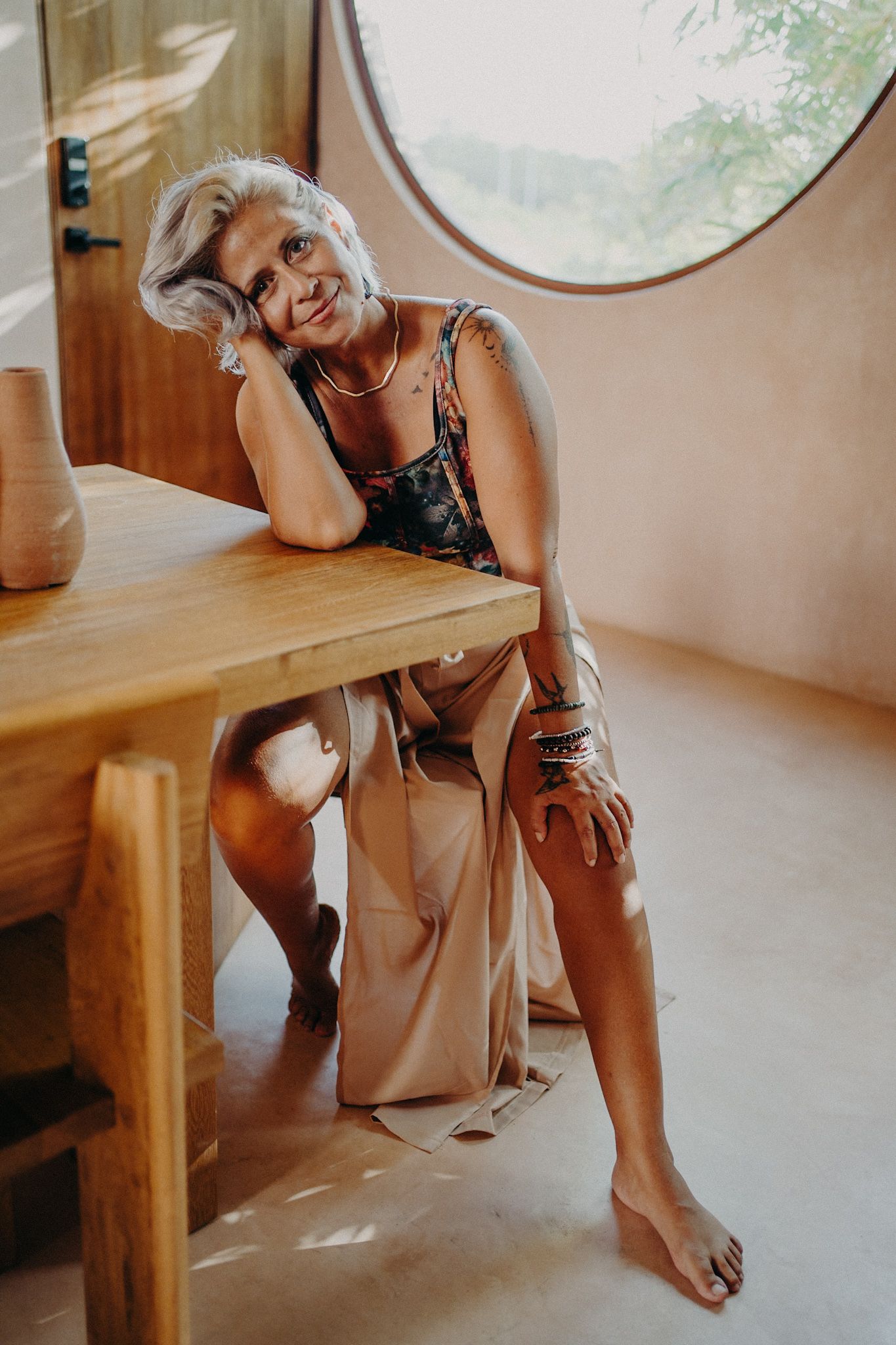 Smiling mature woman with gray hair sitting barefoot by a wooden table in a sunlit boho-style room with a round window.