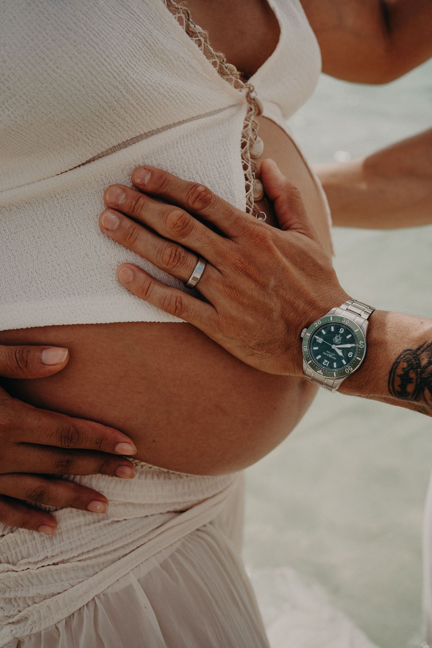 Pregnant woman in white dress with partner's hand on baby bump, wearing green dial watch and tattoo.