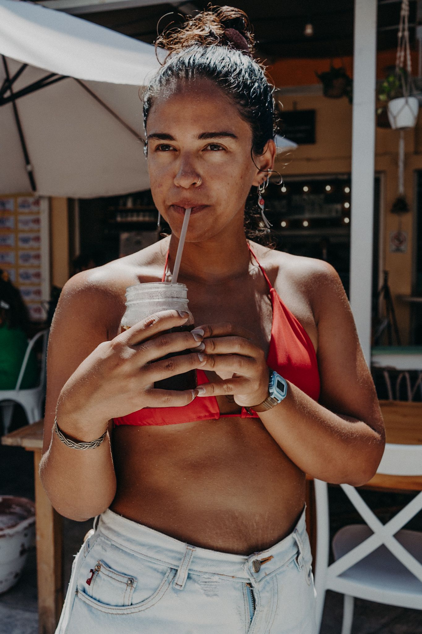 Woman in red bikini top and jeans sipping a drink through a straw at an outdoor cafe on a sunny day.