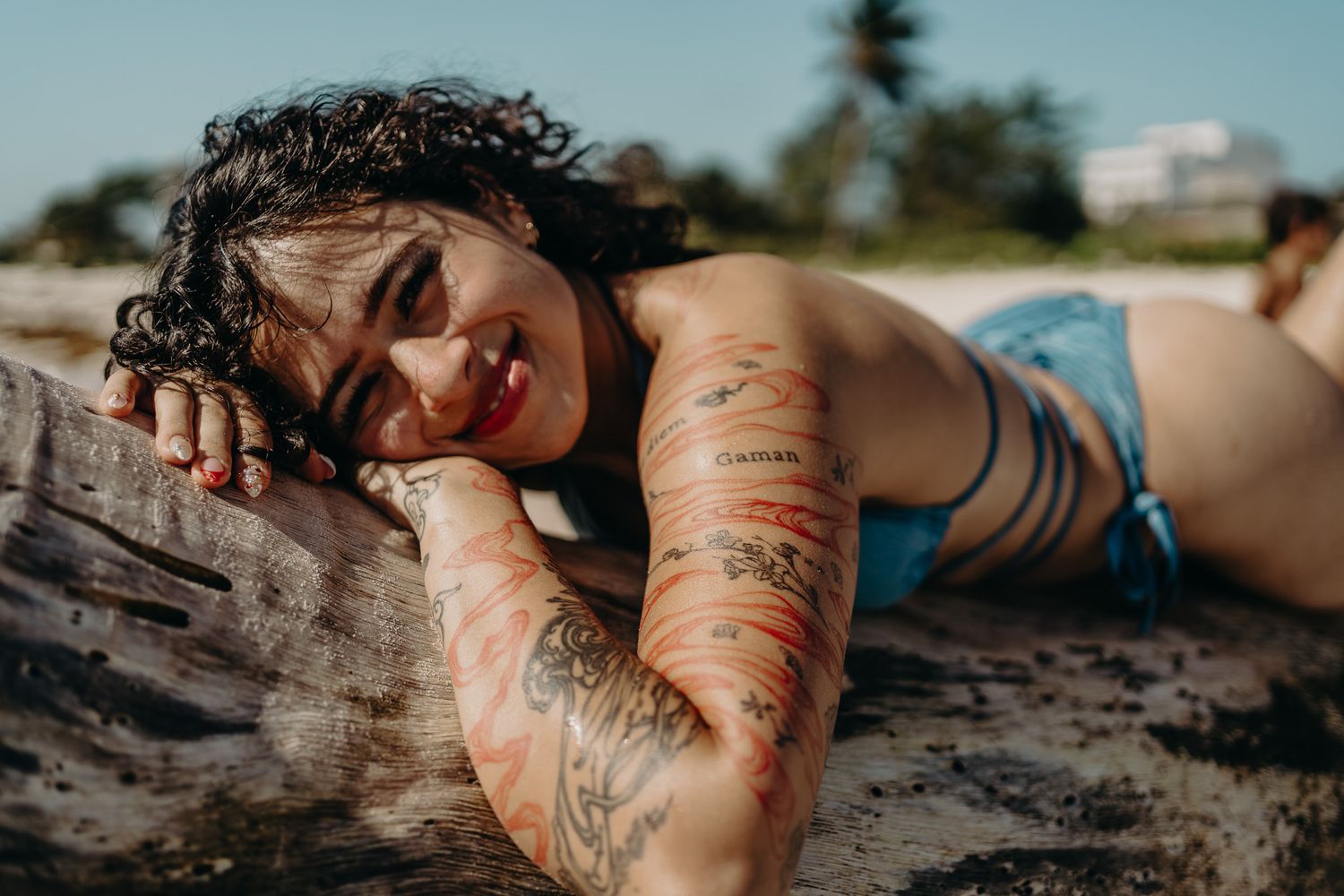 Smiling tattooed woman lying on driftwood at the beach with wet hair and blue bikini top.