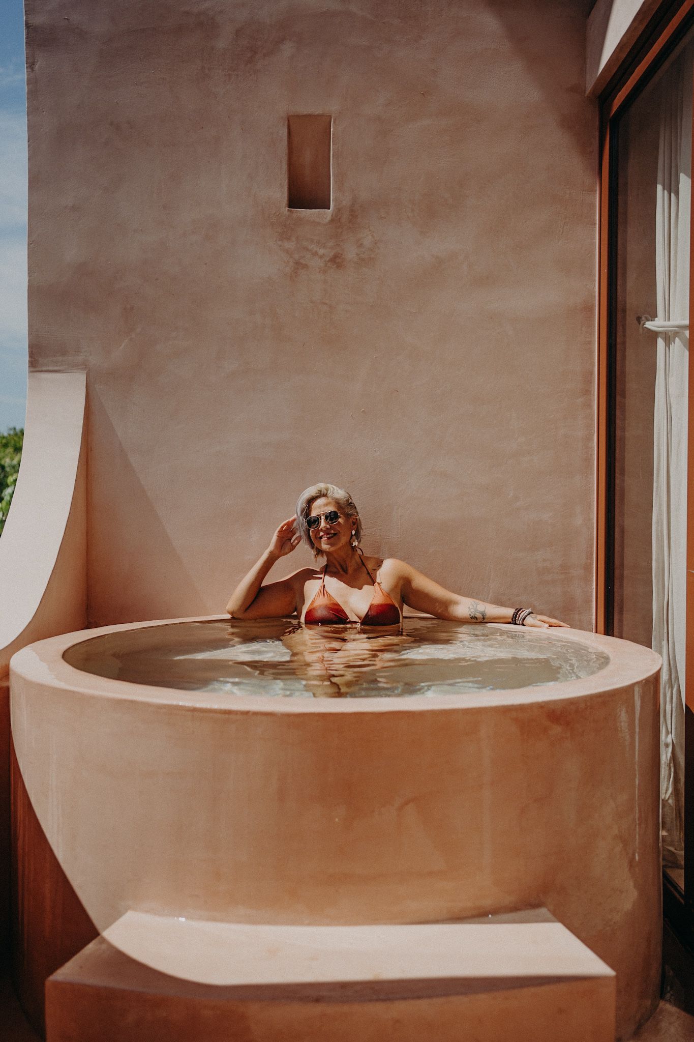 Woman in red bikini and sunglasses relaxing in a round outdoor plunge pool with terracotta walls.