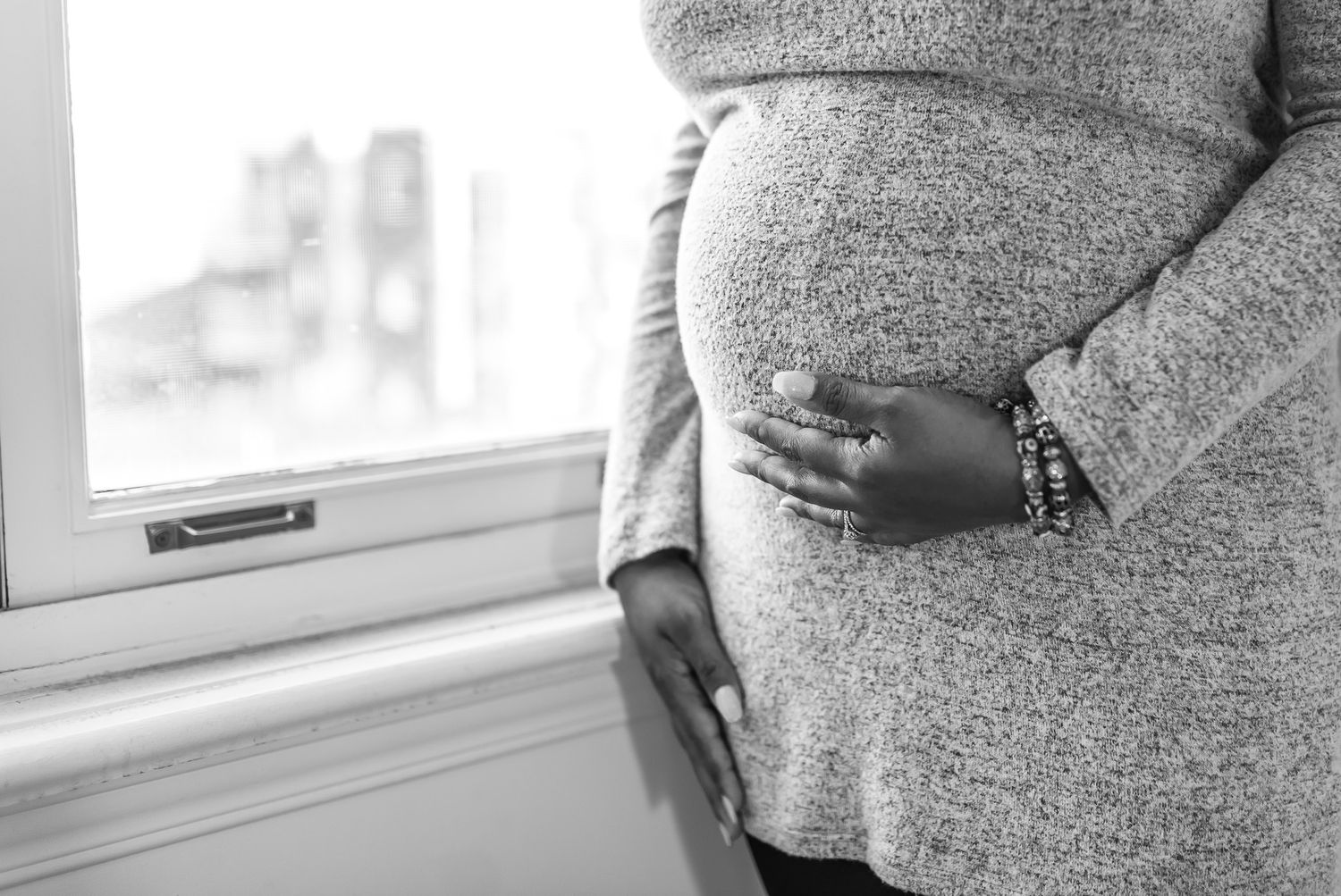 Black and white photo of a pregnant woman cradling her baby bump while standing near a window.