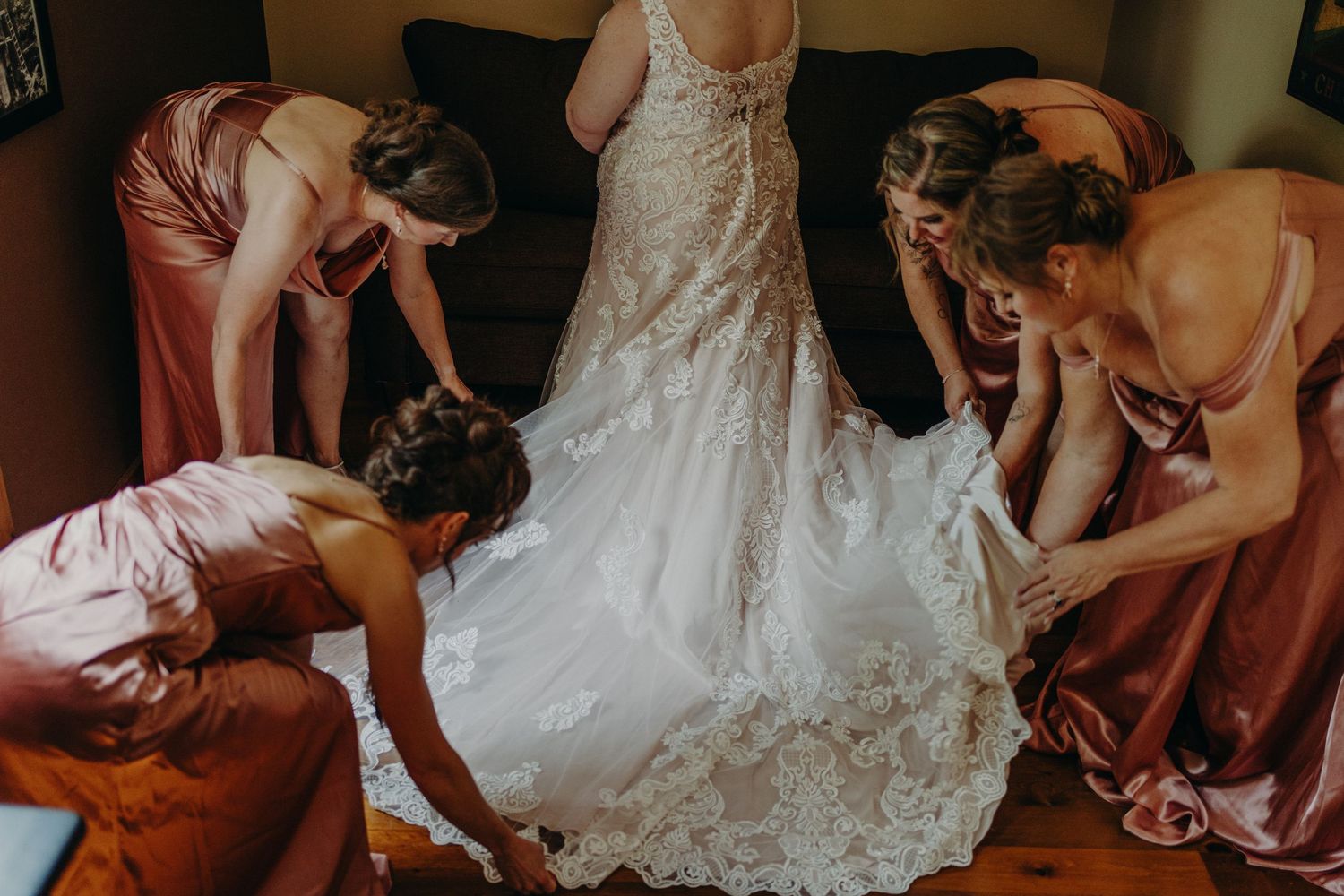 Bridesmaids in dusty rose satin dresses helping bride into lace wedding gown before ceremony.