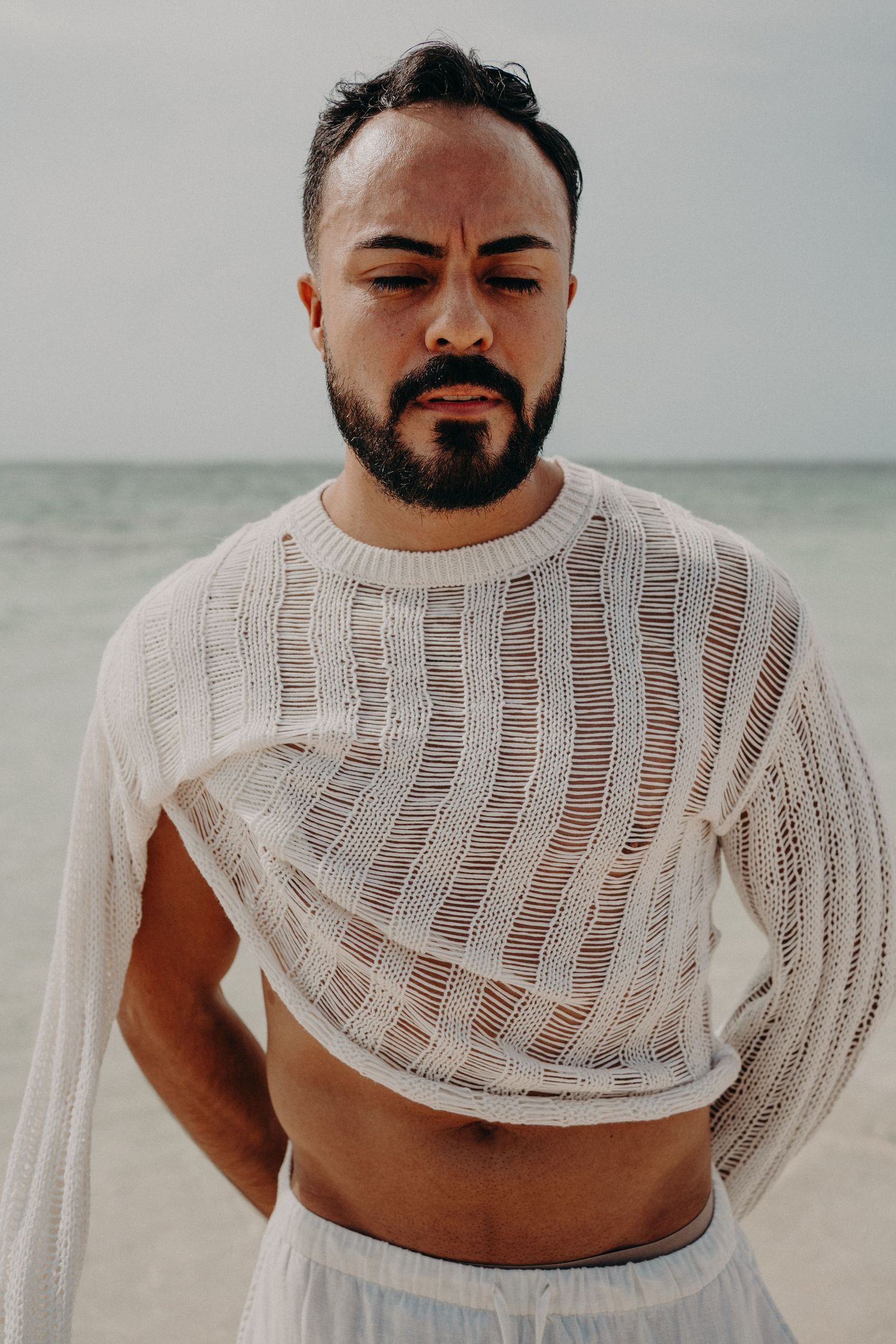 Bearded man in white sheer knit crop sweater and white pants standing on a beach with ocean in background.