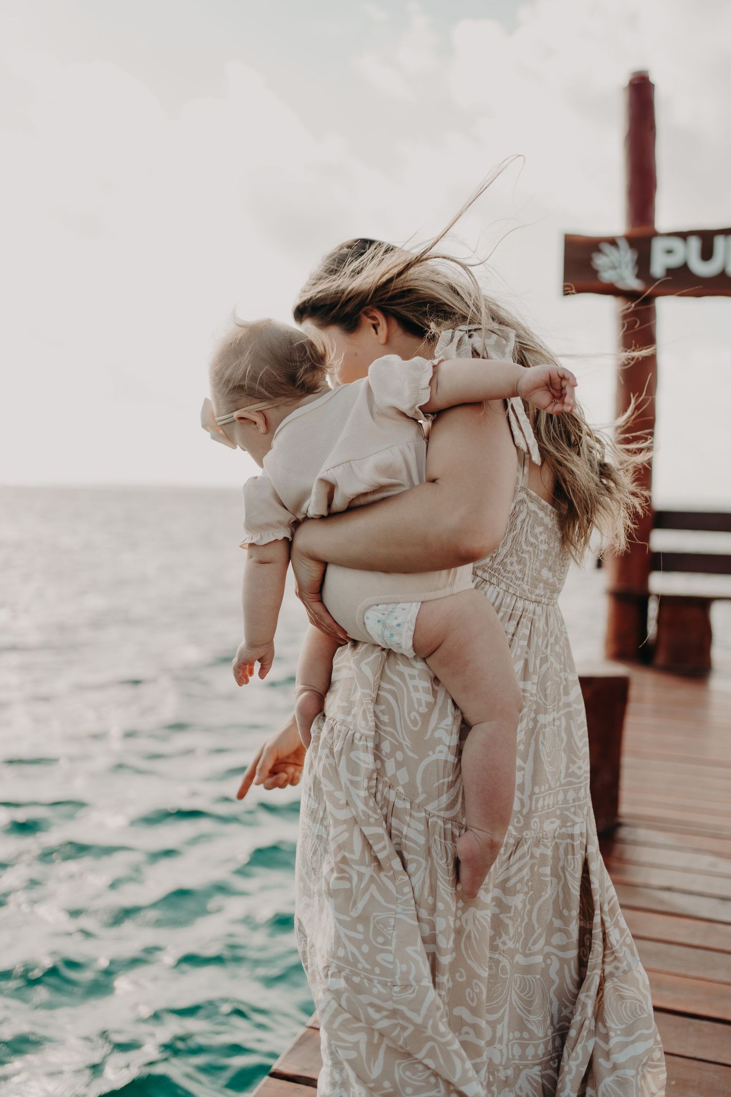 A mother holding her toddler on a wooden pier by the turquoise ocean, both wearing neutral boho outfits.