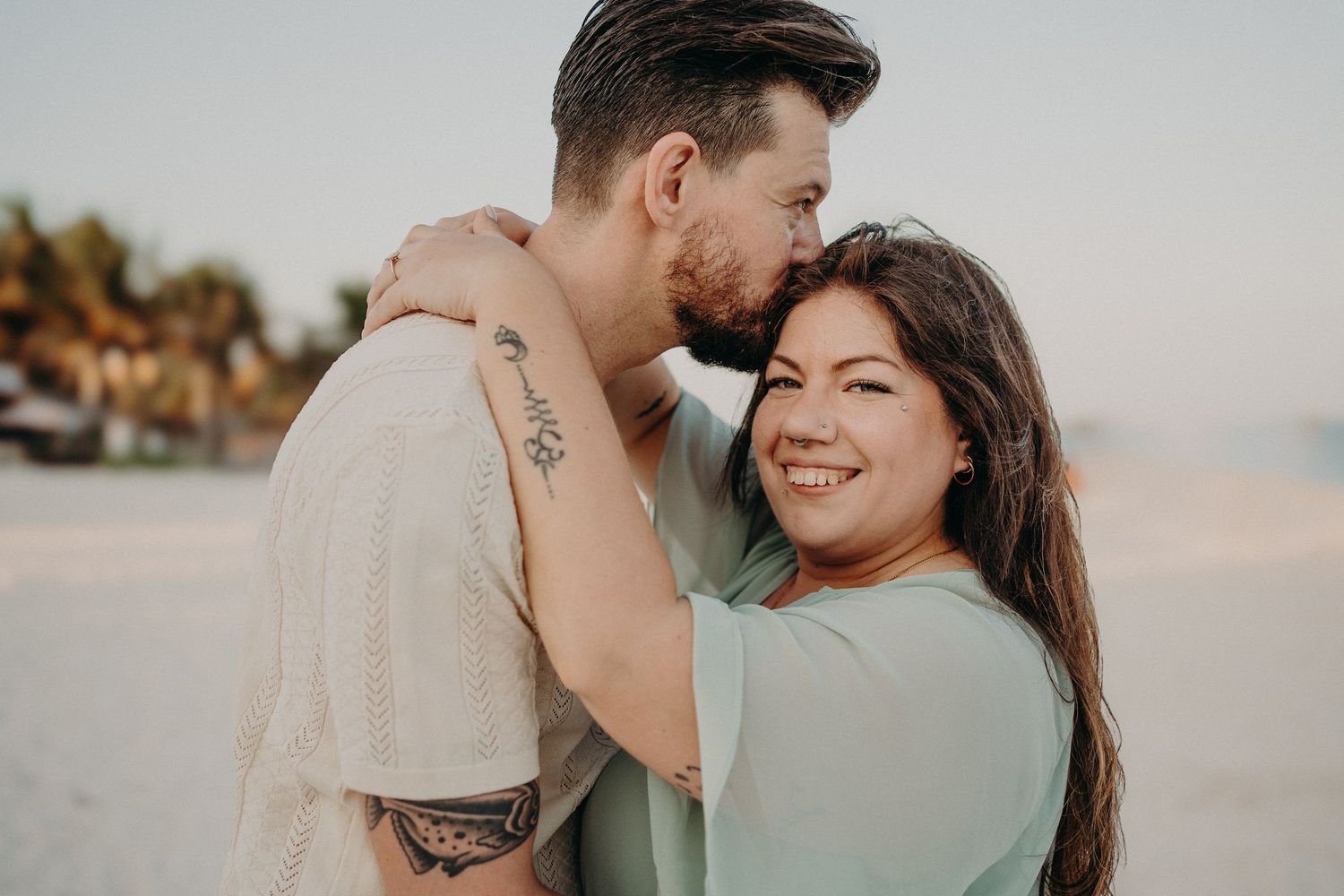 Couple sharing a romantic beach moment, man kissing woman's forehead while she smiles, palm trees in background.