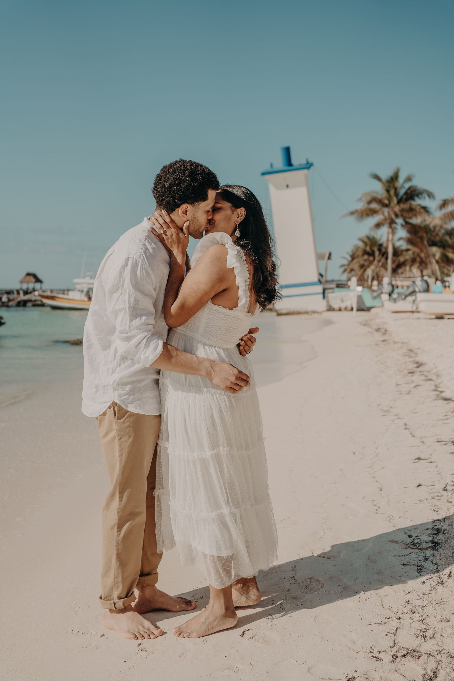 Couple sharing a romantic kiss on a tropical beach, woman in white dress, man in white shirt and khaki pants.