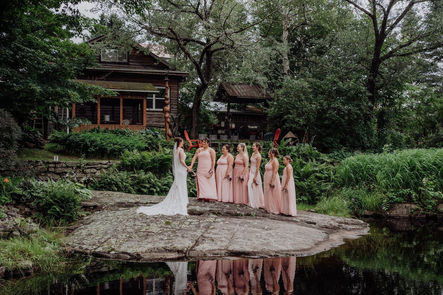 Bridal party in pink gowns posing on rocks beside reflective pond with rustic lodge backdrop.
