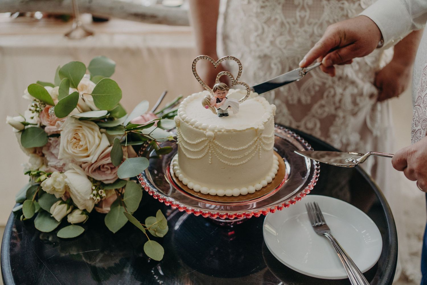 Bride and groom cutting a small white wedding cake decorated with a heart topper and figurines.