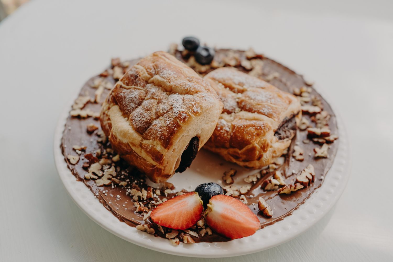 Flaky pastries with blueberry filling on a plate with strawberries, granola, and chocolate sauce.