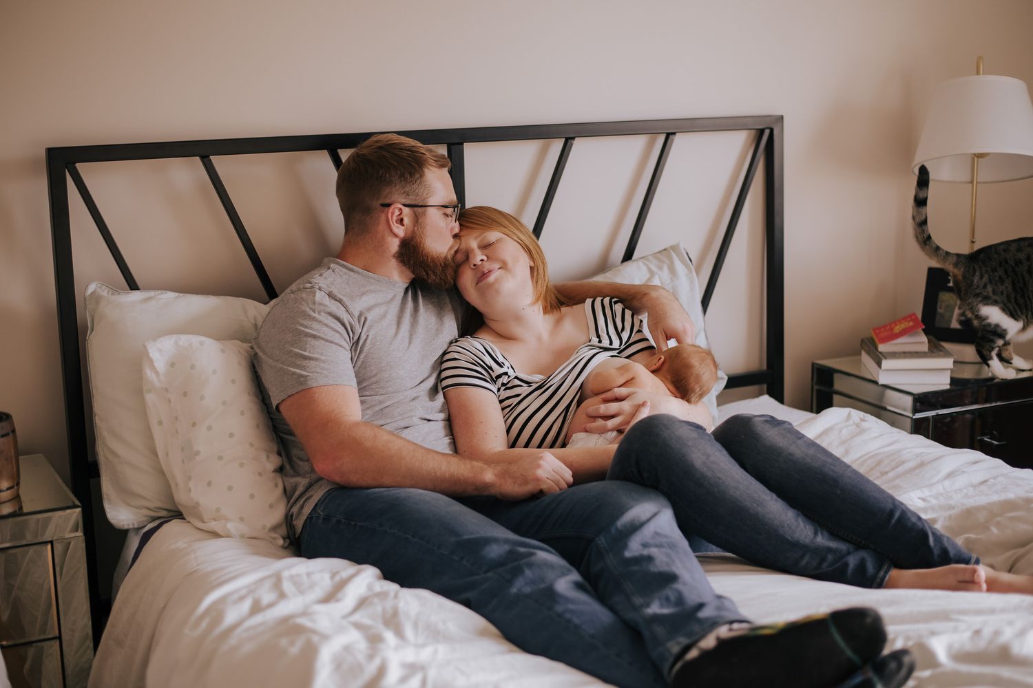 Couple relaxing on bed with newborn baby, sharing tender moment in cozy bedroom with cat nearby.