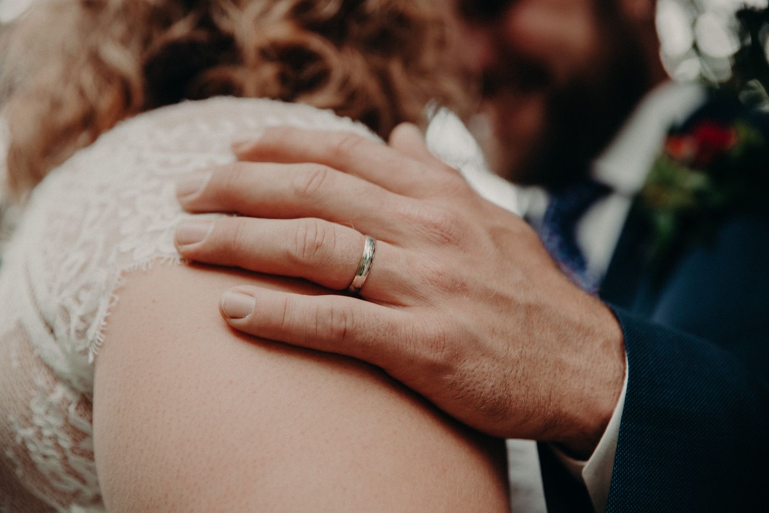 Close-up of groom's hand with wedding band resting on bride's shoulder in lace wedding dress.