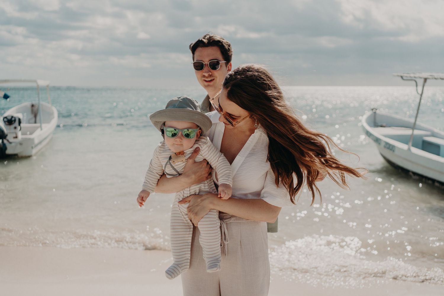 Couple with sunglasses holding a baby on a sunny beach with boats and turquoise water in the background.