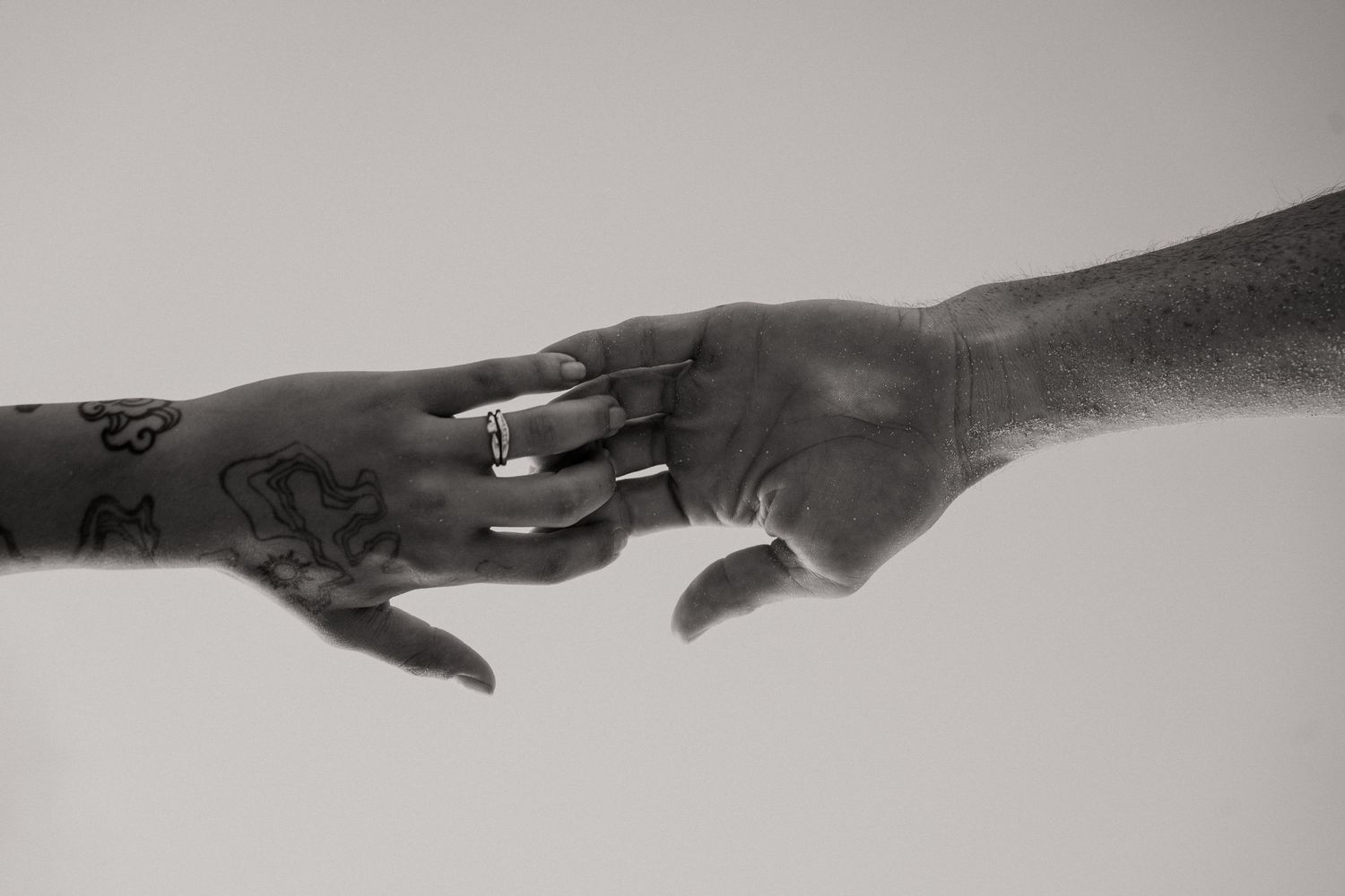 Black and white photo of two hands touching fingertips, one with tattoos and rings, symbolizing connection.