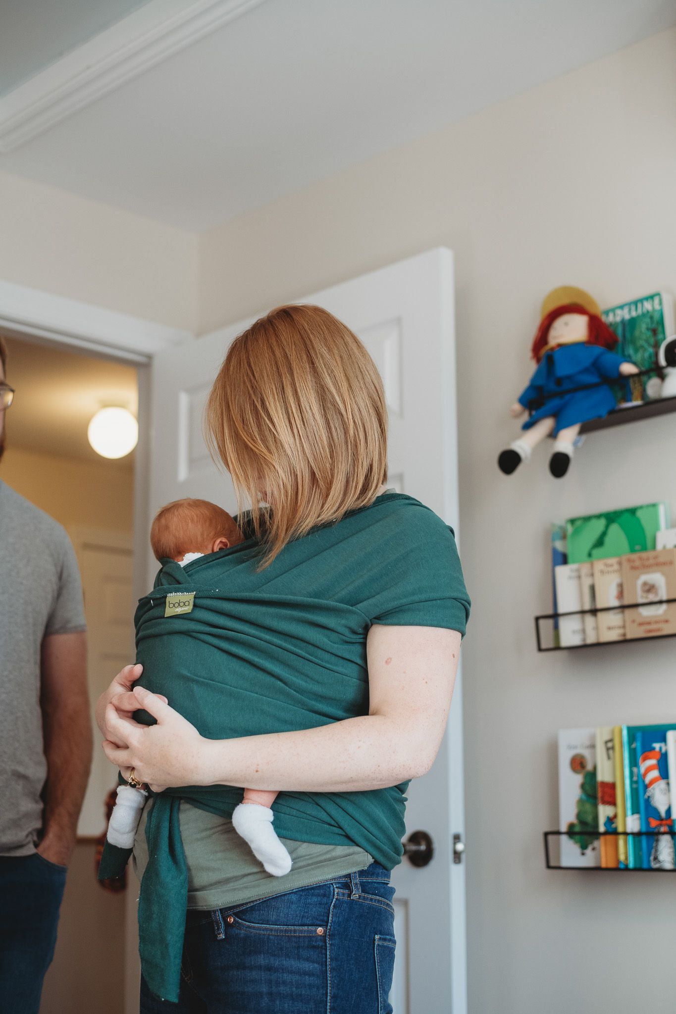 Mother in green shirt carrying newborn baby in wrap carrier inside a nursery with book shelves and Madeline doll.