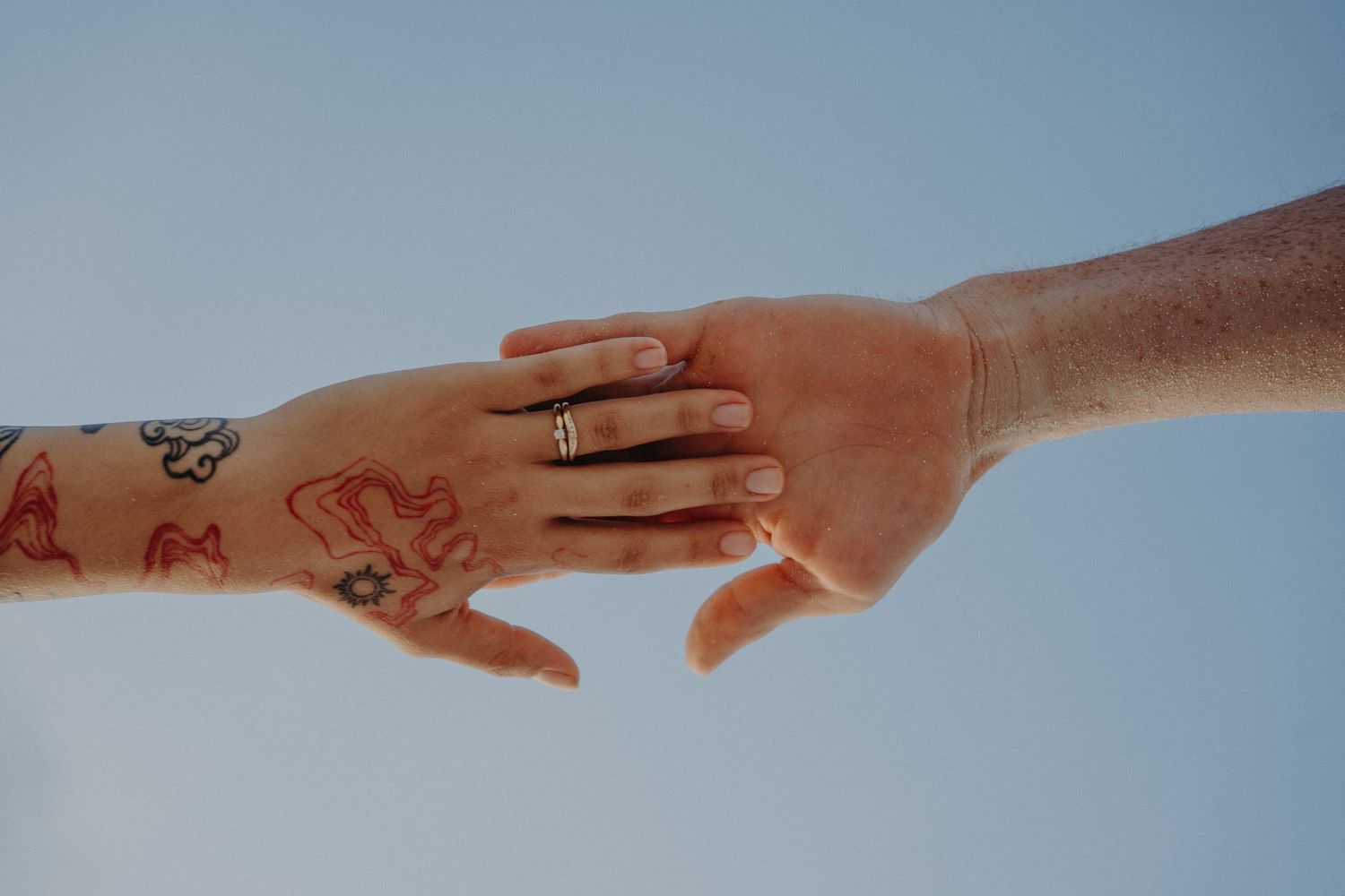 Two hands touching, one with colorful tattoos and a gold ring, against a soft blue sky background.