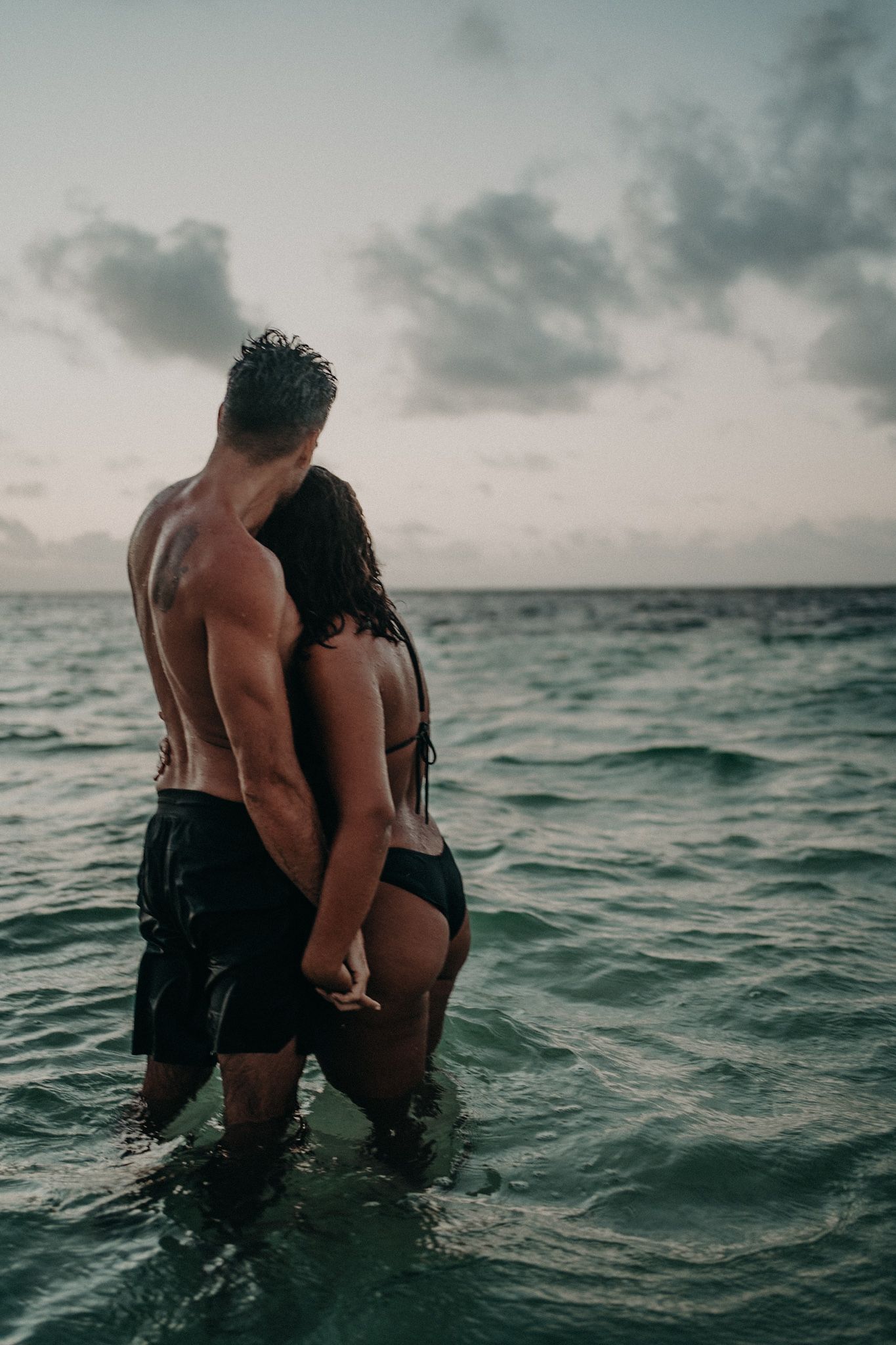 Romantic couple standing together in shallow ocean water at dusk, man shirtless in swim trunks, woman in black bikini.