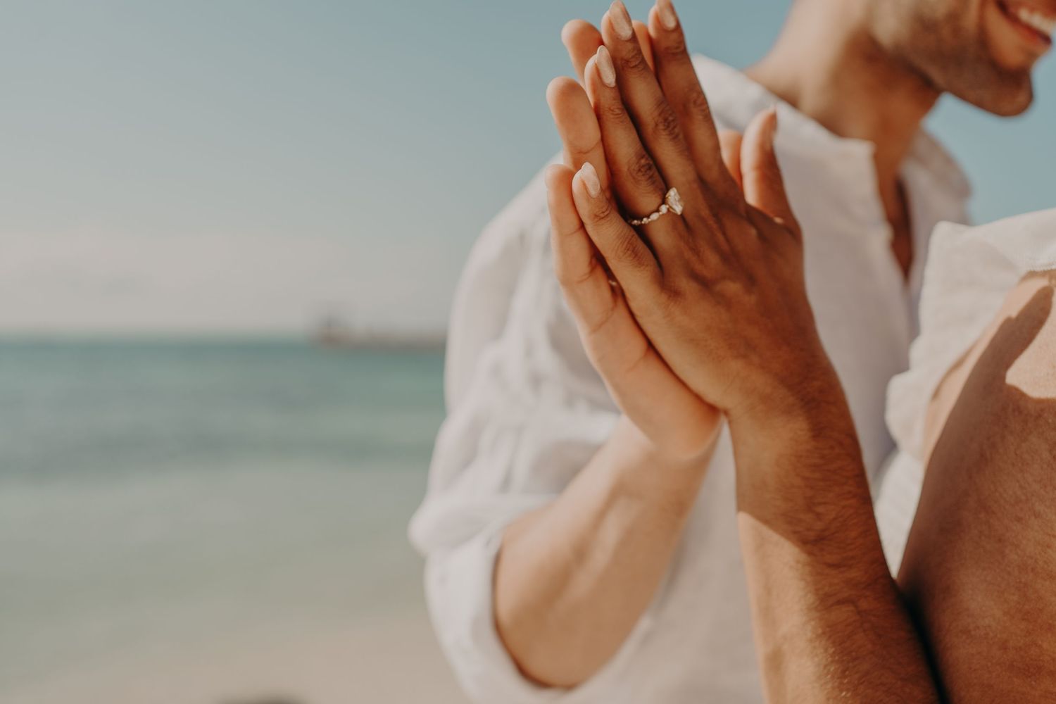 Couple holding hands on beach showing gold engagement ring, ocean in background.