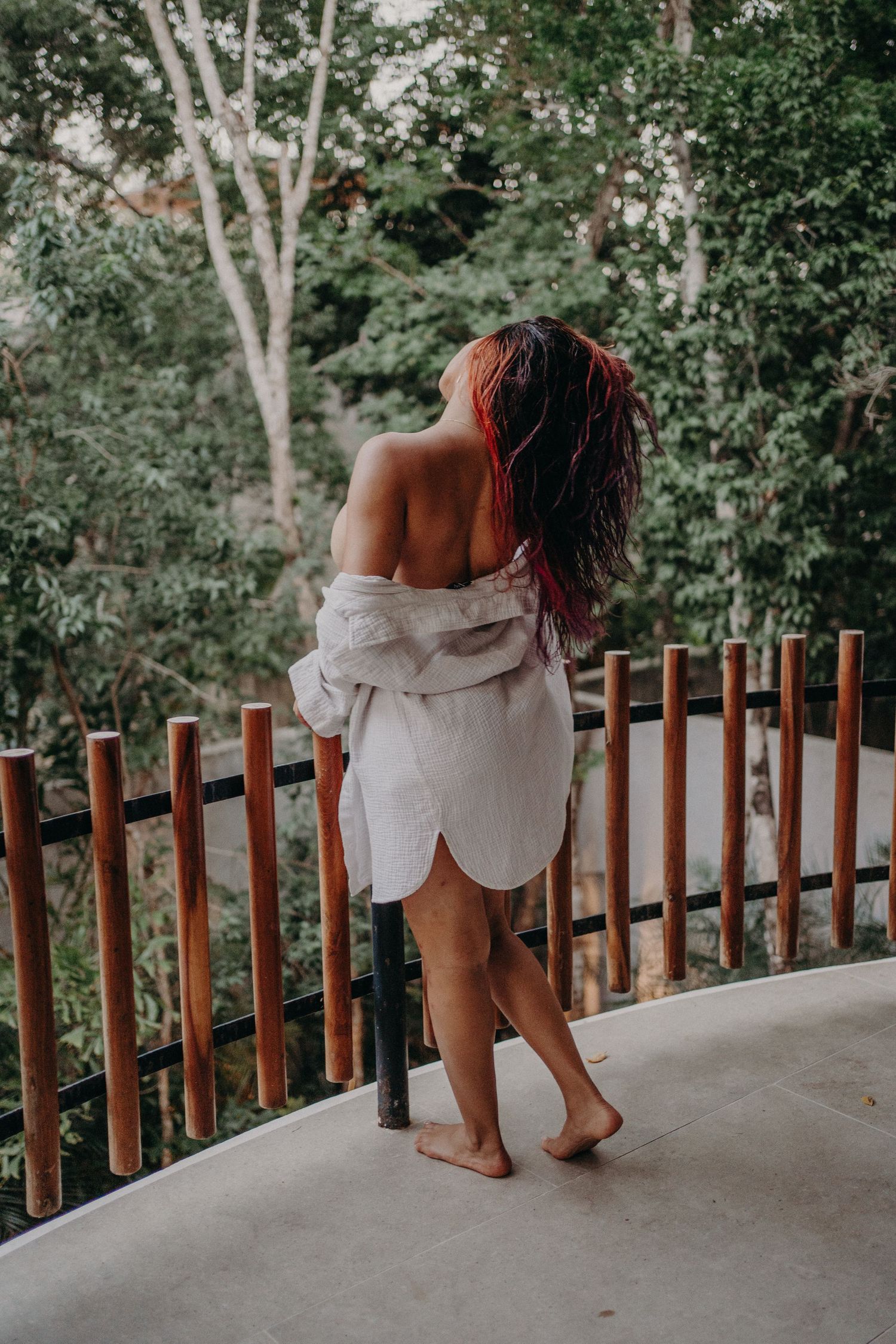 Woman in white off-shoulder shirt stands barefoot on balcony overlooking lush green tropical forest.