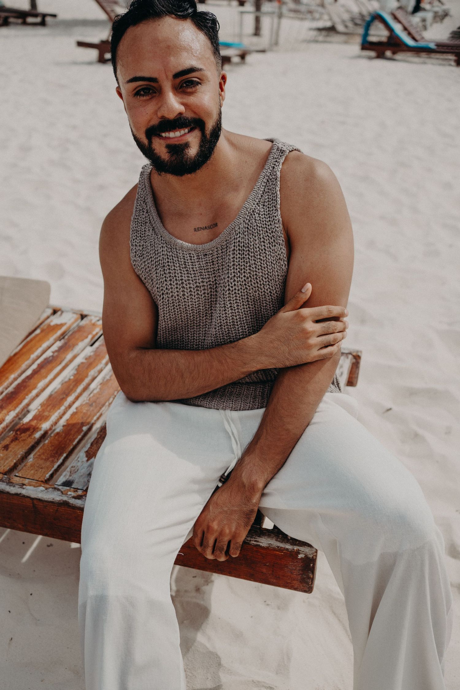 Smiling bearded man in gray knit tank top and white pants sitting on weathered wooden bench on sandy beach.