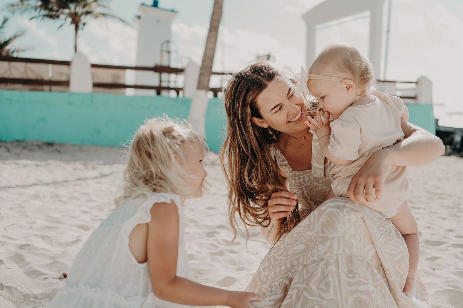 A mother sits on a sandy beach smiling and playing with her two young children on a sunny day.