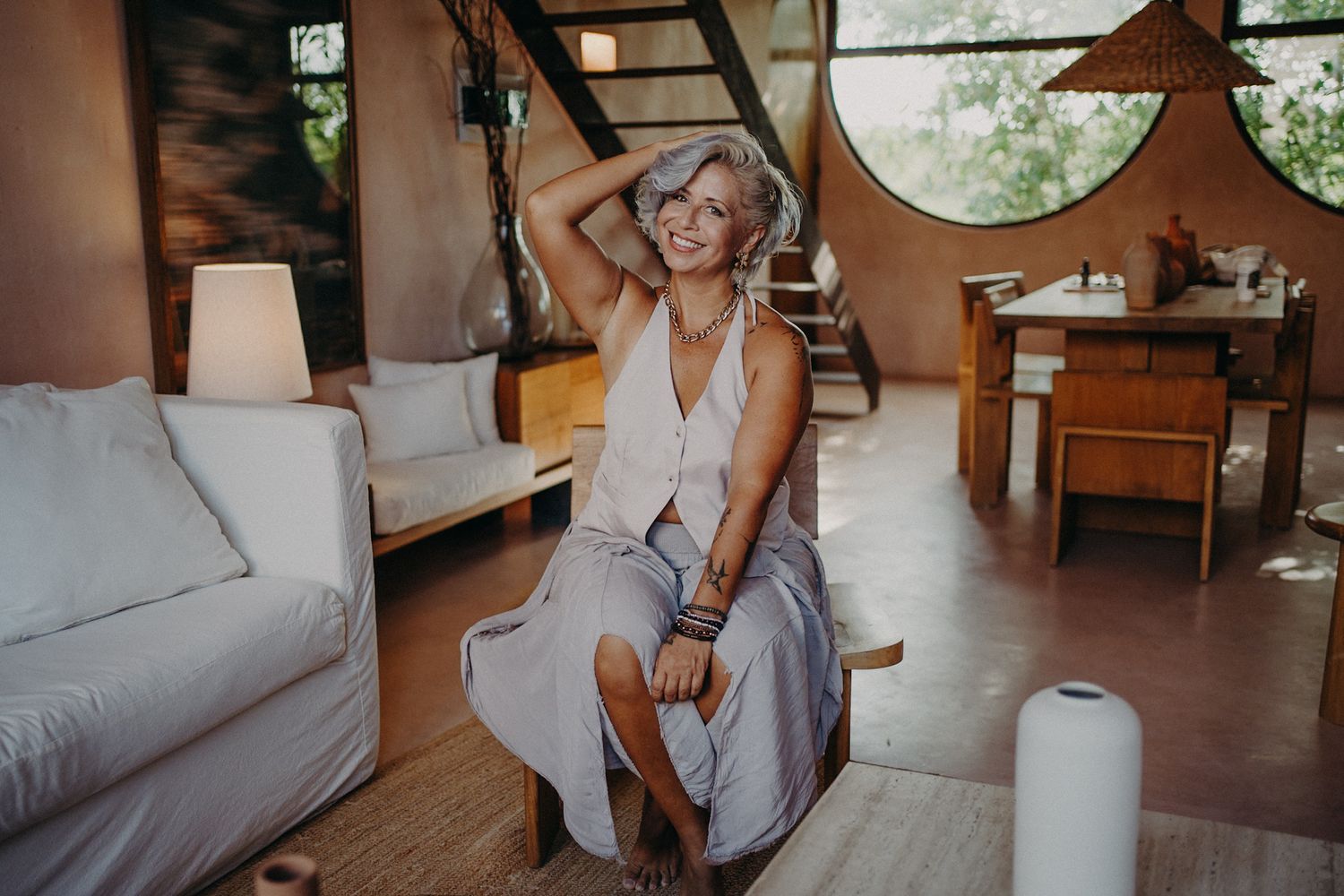 Smiling mature woman with short gray hair sitting relaxed in a modern rustic living room with natural light.