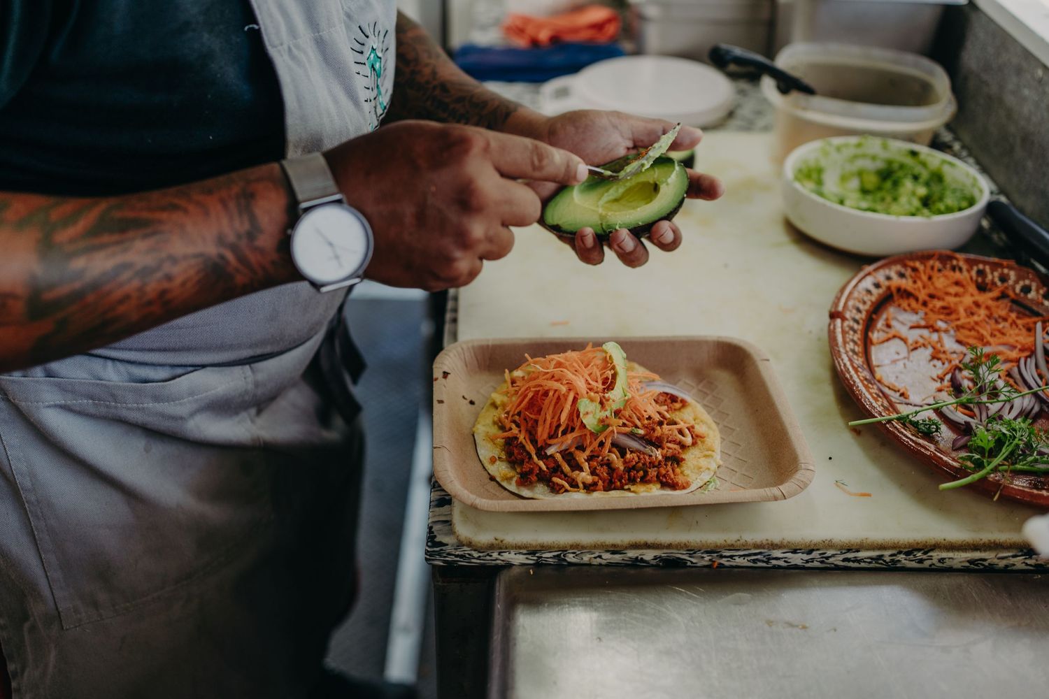 Tattooed chef slicing avocado onto a taco with shredded meat on a wooden board in a restaurant kitchen.