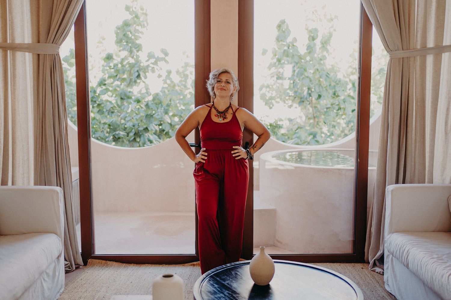 Woman in red jumpsuit standing confidently in elegant room with large glass doors and lush greenery outside.