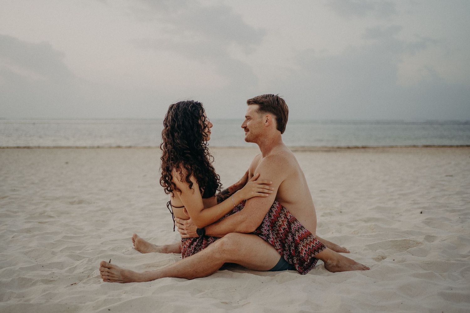 Couple sitting together on sandy beach, sharing a romantic moment by the ocean on an overcast day.