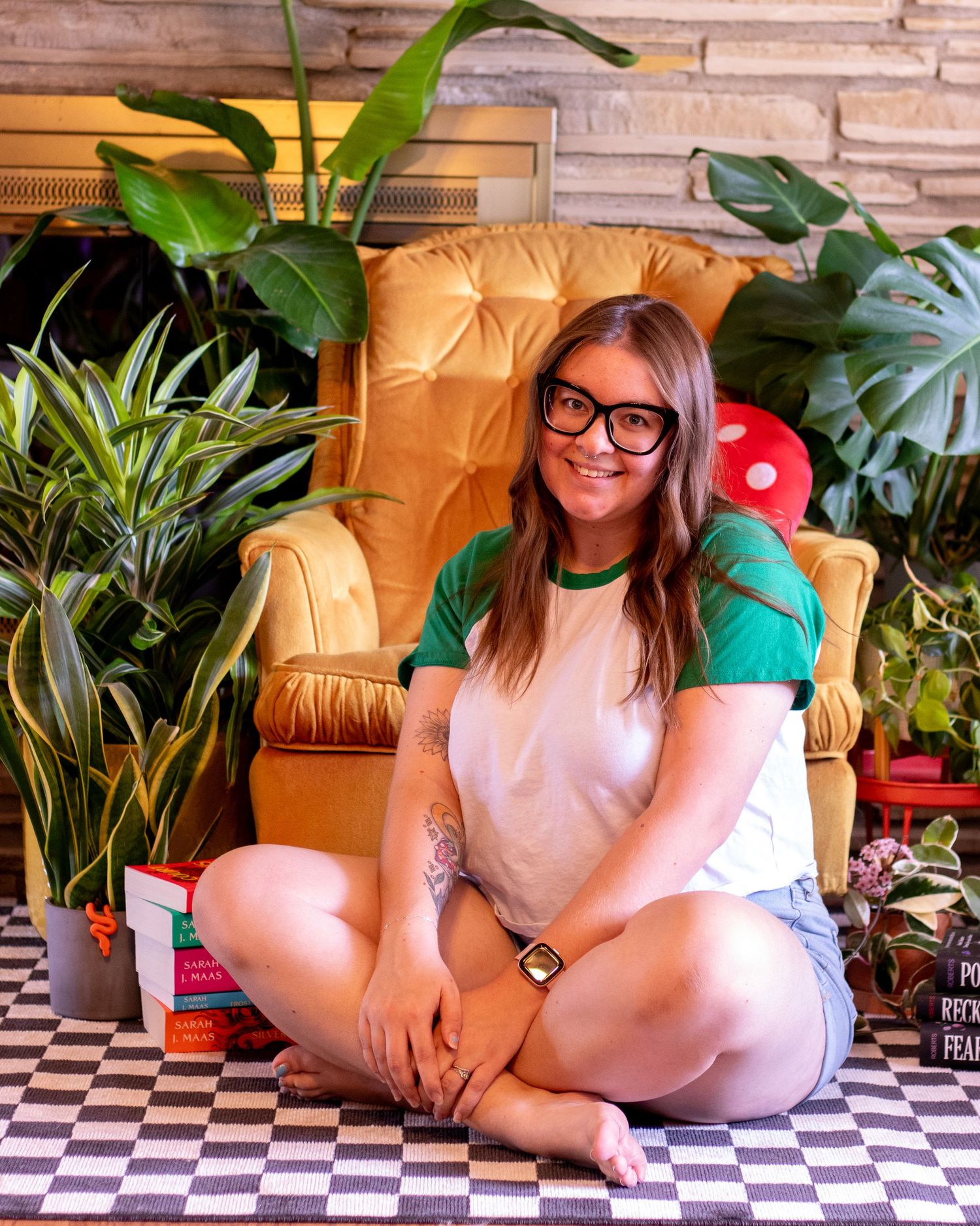 A girl sitting in front of a vintage yellow chair surrounded by books and plants.