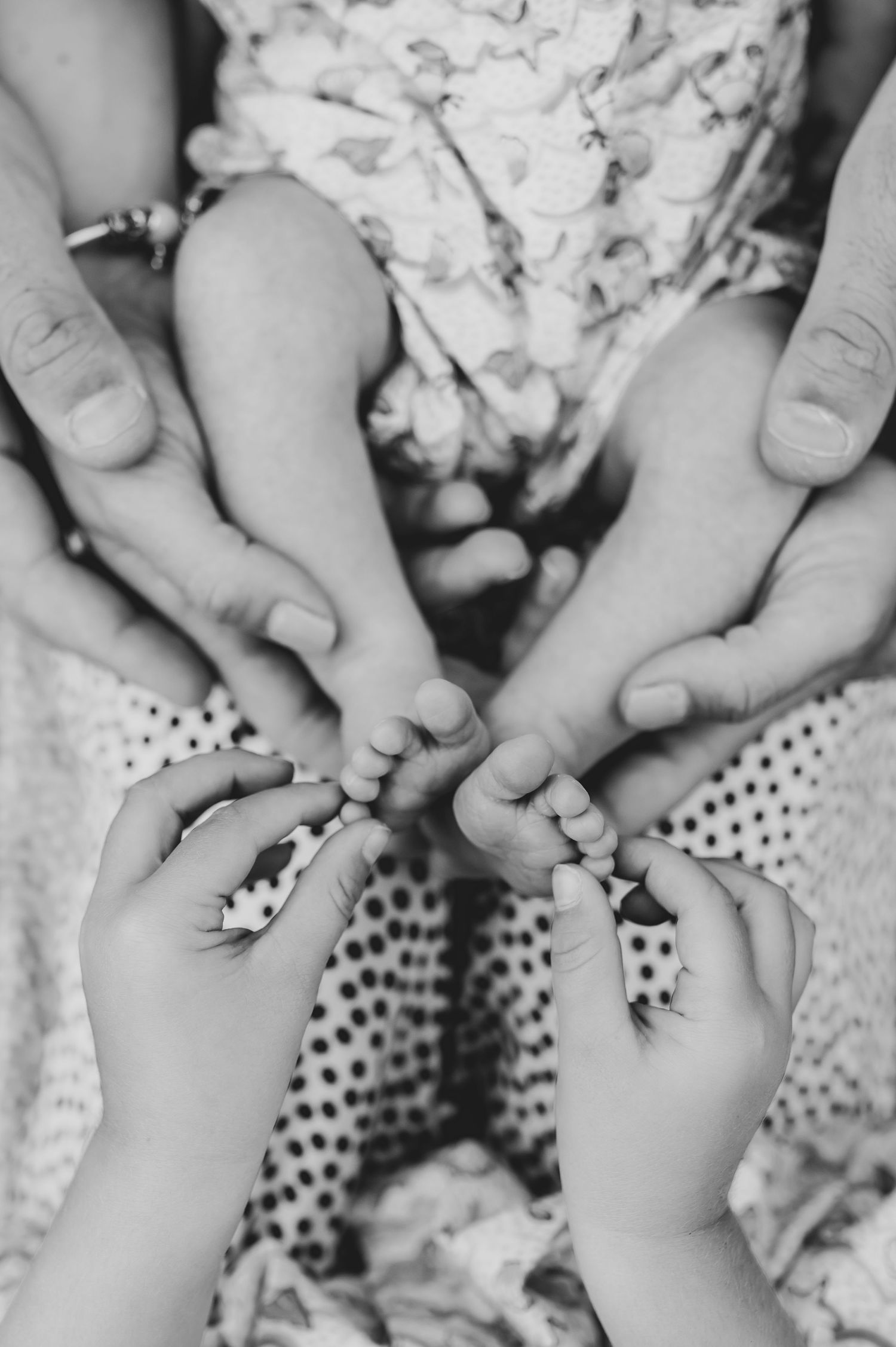 Black and white close-up of tiny baby feet held by multiple hands against patterned fabric.