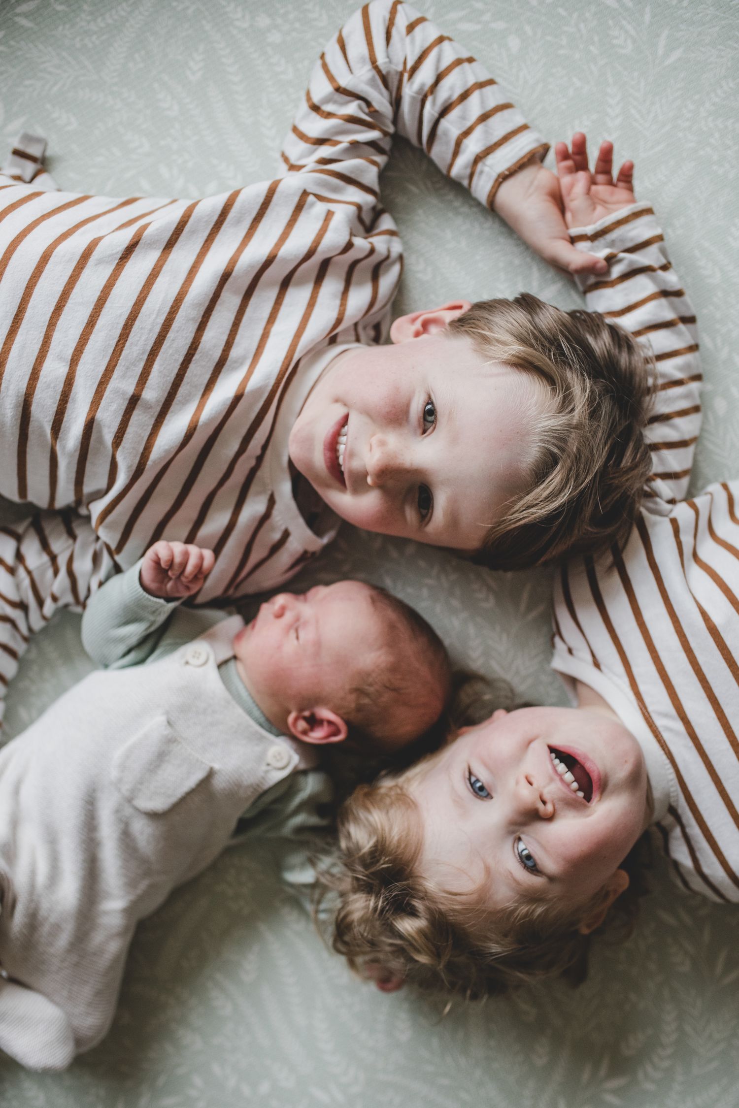 Three siblings lying together on gray fabric with bright smiles and striped clothing.