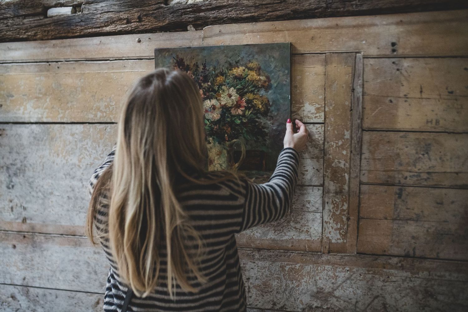 Woman in striped shirt admiring floral artwork hanging on rustic wooden wall.